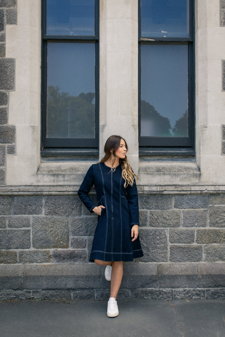 A woman wearing the Vassalli Sienna Dress in Ink Denim with white sneakers stands before a stone wall with two tall windows, looking to her right and resting her hands by her sides.