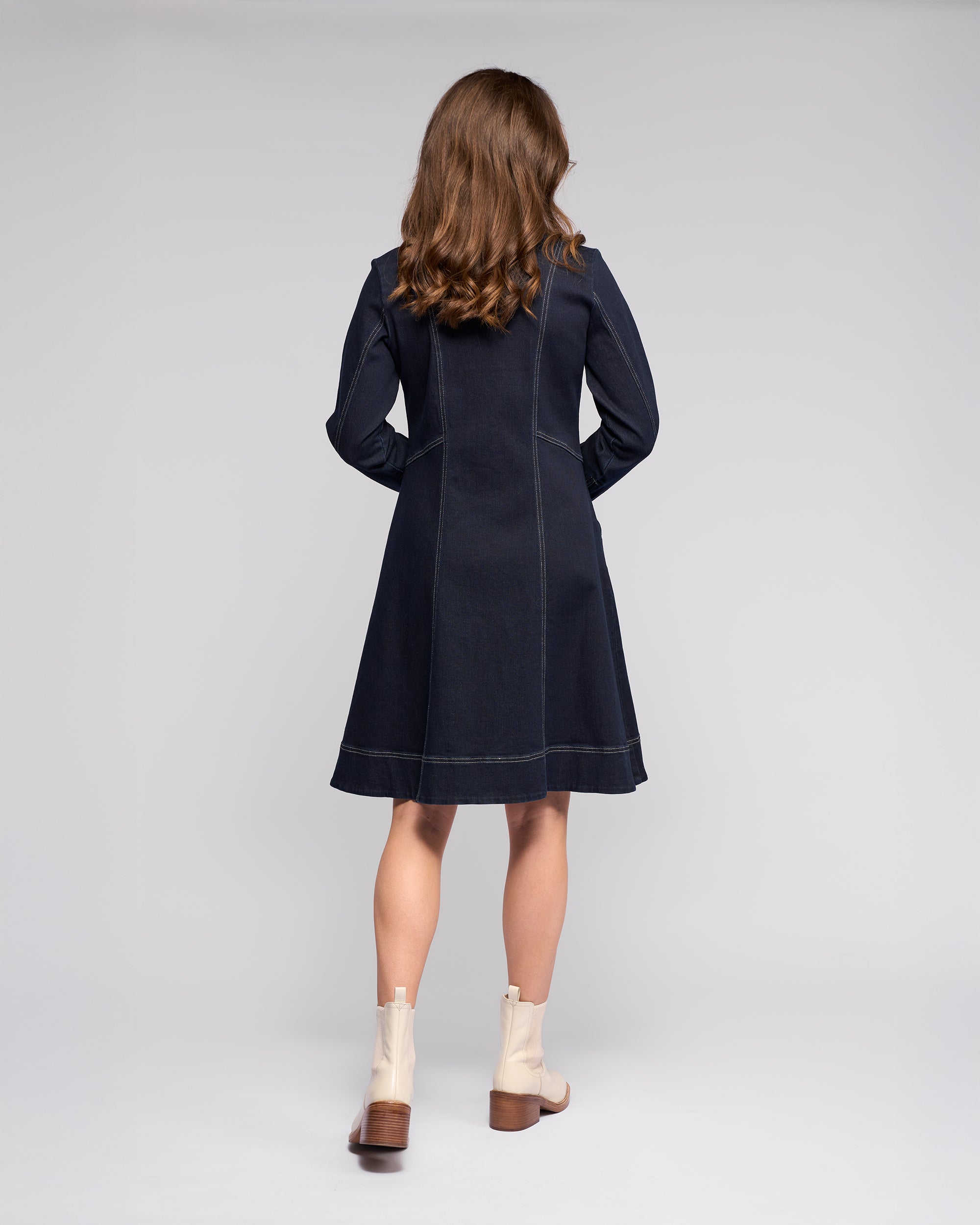 A woman with wavy brown hair faces away, wearing the Vassalli Sienna Dress in Ink Denim with light beige ankle boots against a plain light background.