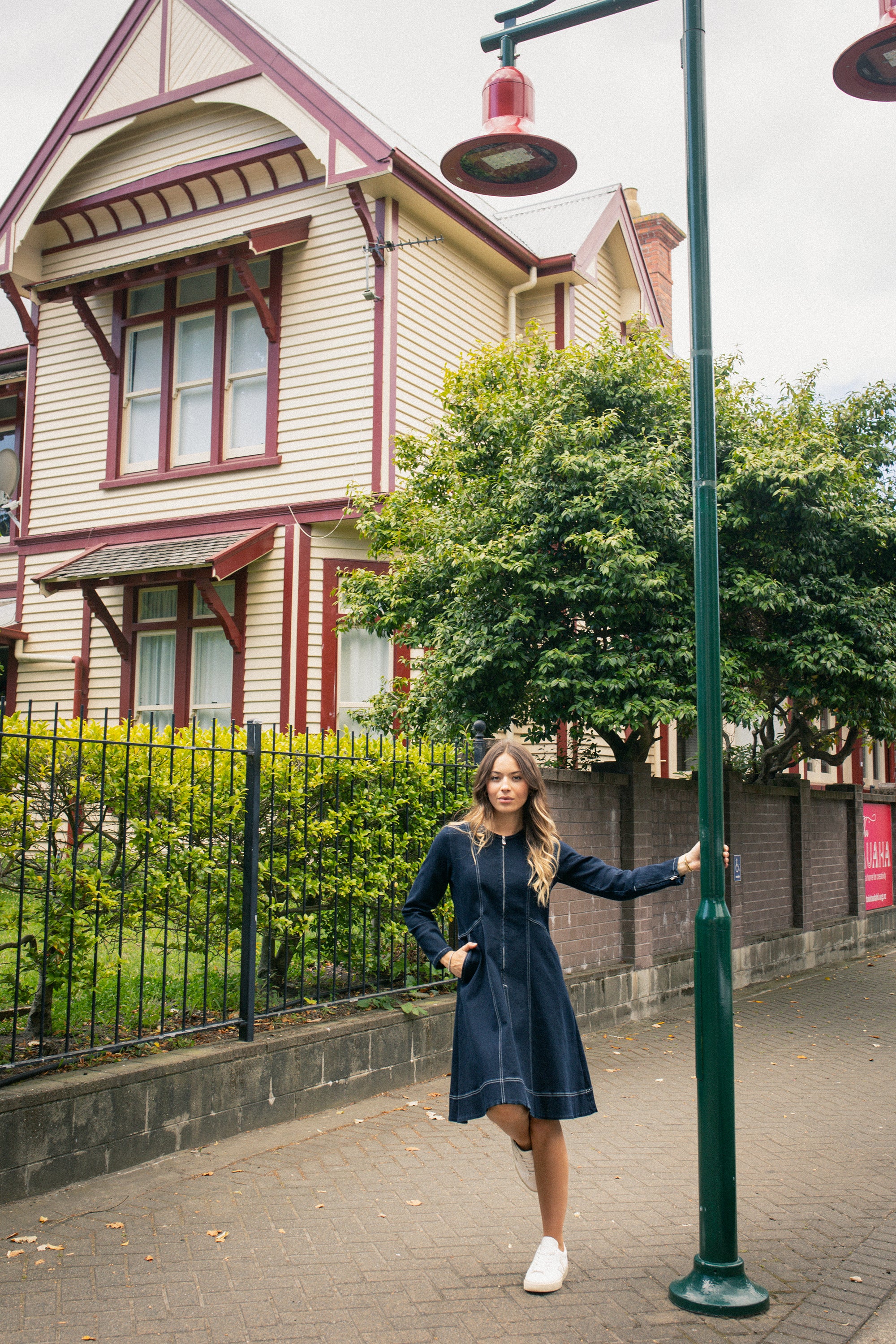 A woman wearing the Vassalli Sienna Dress in Ink Denim and white sneakers stands by a green street lamp on a sidewalk, with a large cream and red Victorian-style house, garden, and iron fence in the background.