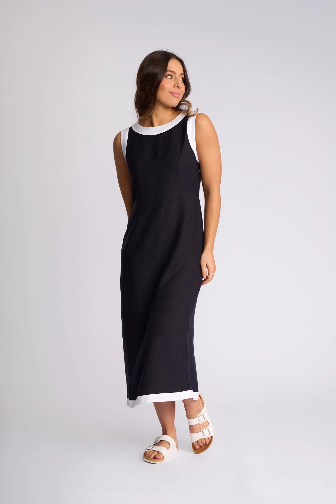 A woman models the Vassalli Oak Dress in Ink/White—an ankle-length, sleeveless summer dress featuring elegant white trim at the neck, armholes, and hem—stylishly paired with white sandals. She stands against a plain white background, looking to the side with a slight smile.