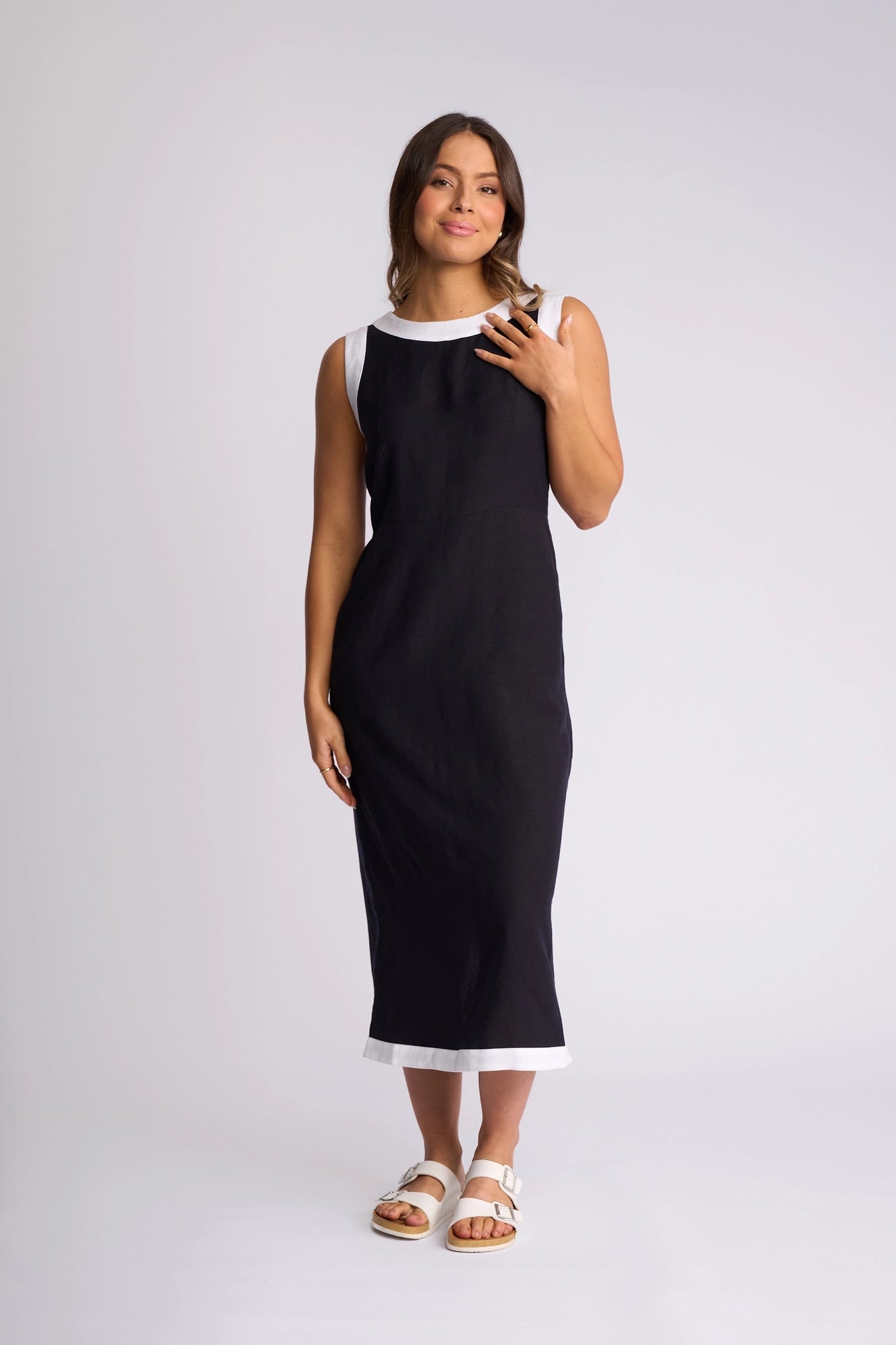 A woman wearing a Vassalli Oak Dress in Ink/White—an ankle-length, sleeveless summer dress with white trim at the neckline and hem—paired with white sandals, standing against a plain light background.