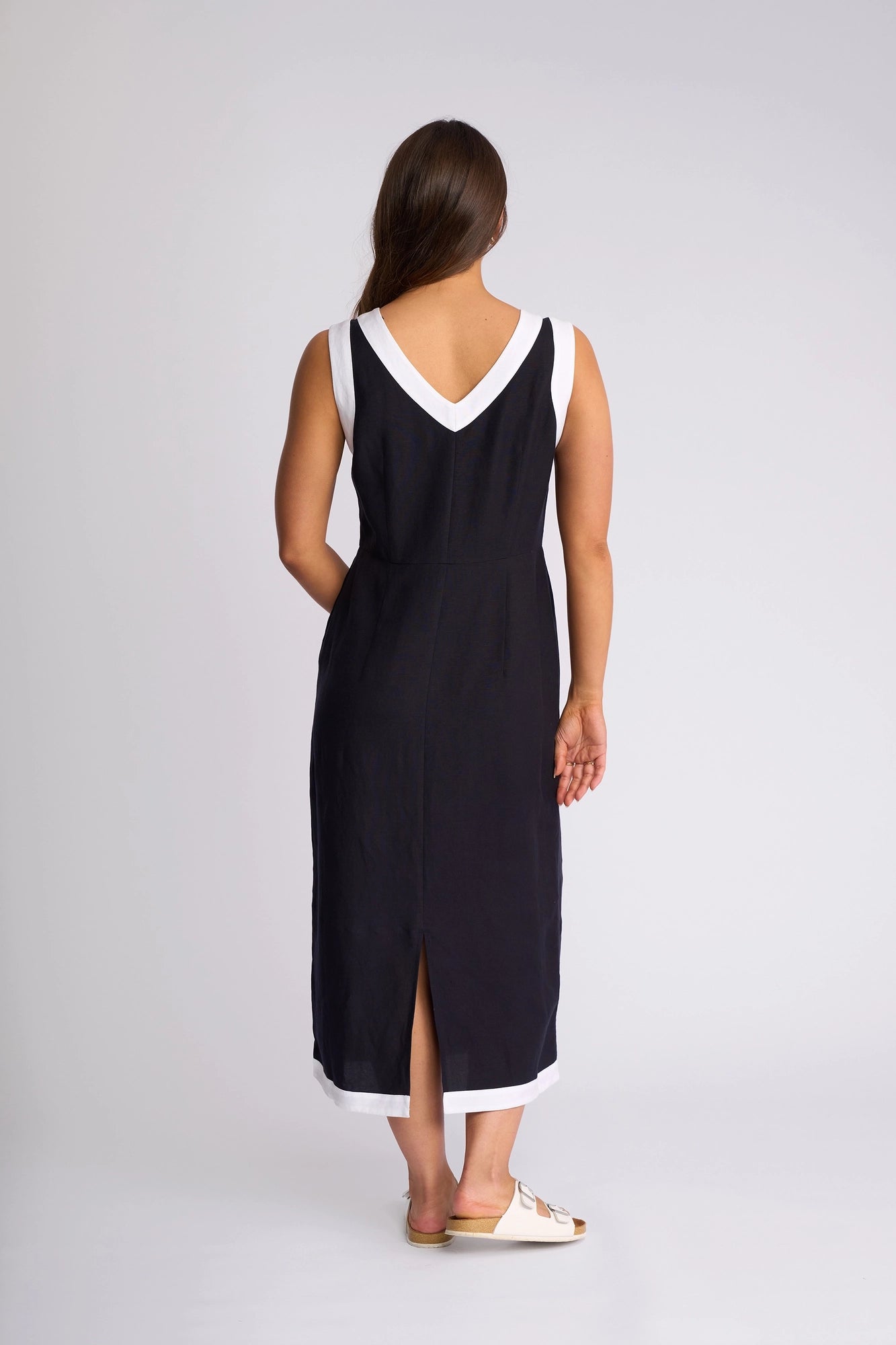 A woman with long brown hair stands facing away against a plain light background, wearing the Vassalli Oak Dress in Ink/White—a sleeveless linen-viscose midi dress featuring white trim and a V-shaped back—and paired with white sandals.