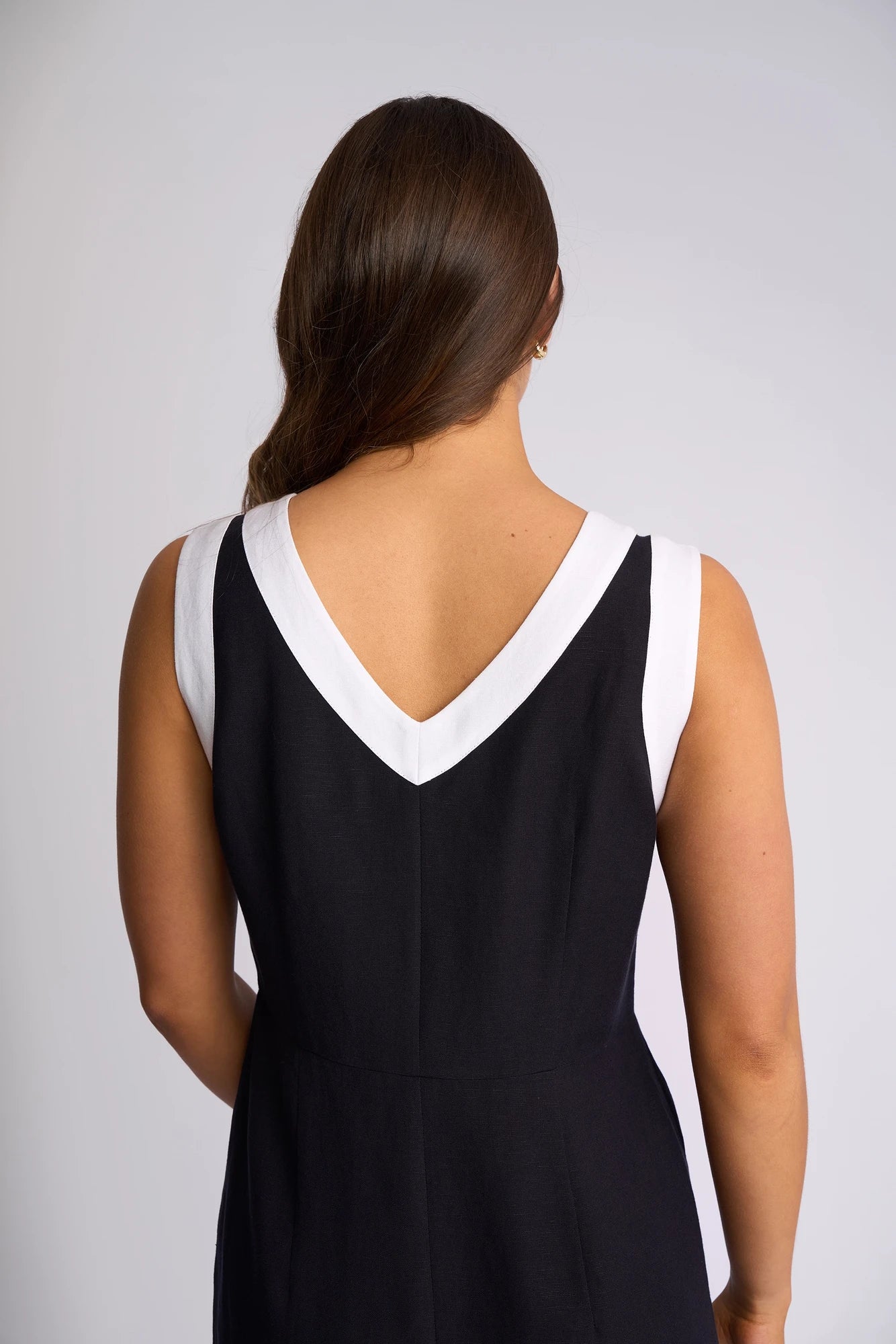 A woman with long brown hair poses with her back to the camera, wearing the Vassalli Oak Dress in Ink/White, a sleeveless summer dress featuring a white V-shaped neckline detail.