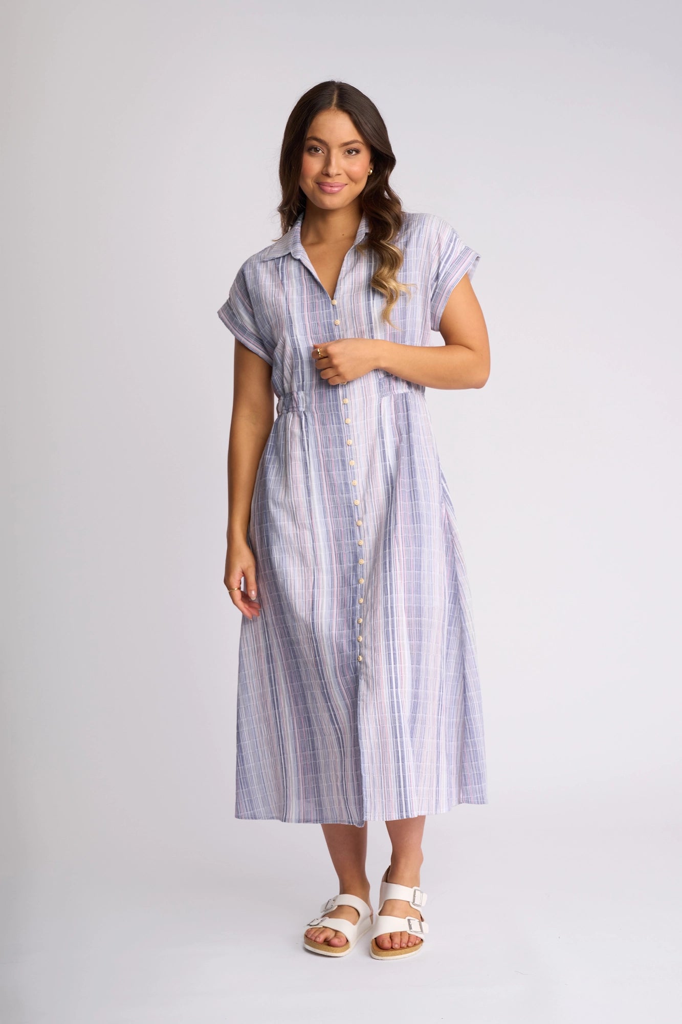 A woman wears the Vassalli Noa Dress - Blue Stripe—a short-sleeve, button-down midi dress in breathable cotton with a smart collar—paired with white sandals and standing against a plain white background.
