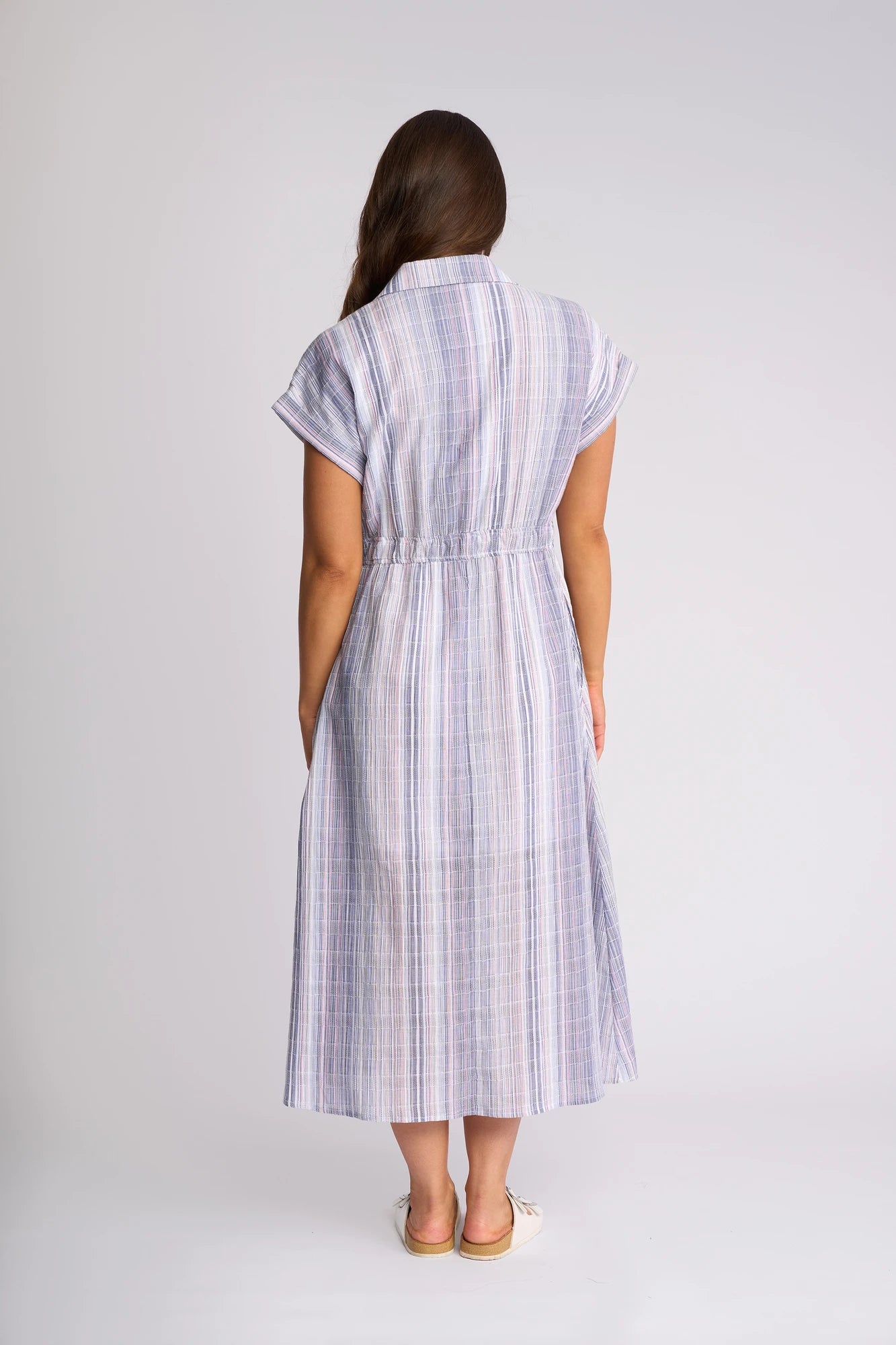 A woman with long brown hair wears the Vassalli Noa Dress – Blue Stripe, a short-sleeve, ankle-length dress made from breathable cotton, paired with sandals, as she stands with her back to the camera against a plain background.