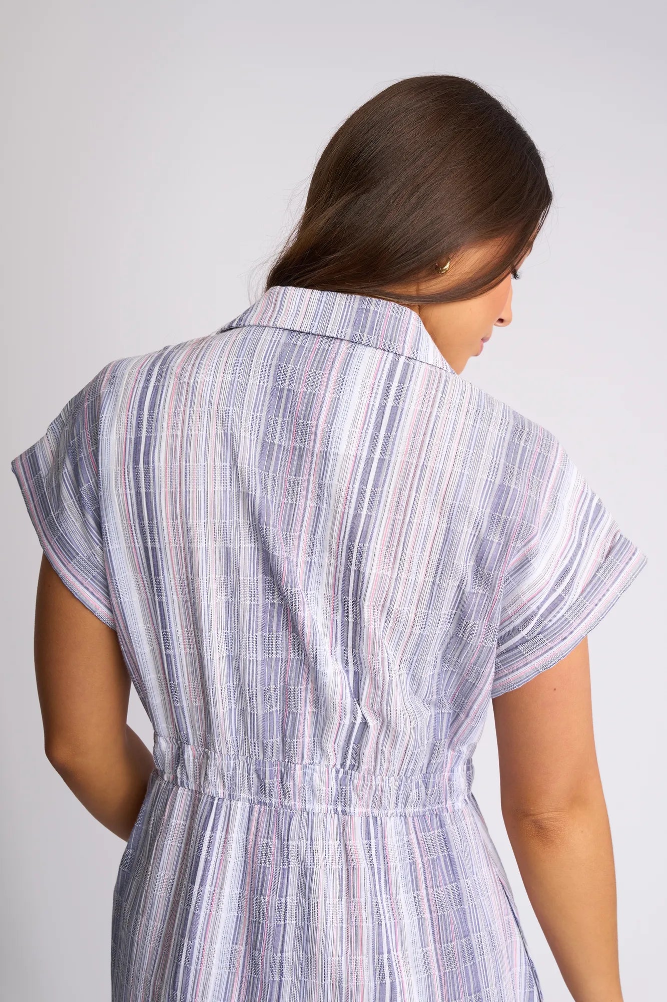 A woman with long brown hair, viewed from behind, is wearing the Vassalli Noa Dress - Blue Stripe—a short-sleeved, collared dress made from breathable cotton with pastel vertical blue stripes and an elastic waist—standing against a plain light background.