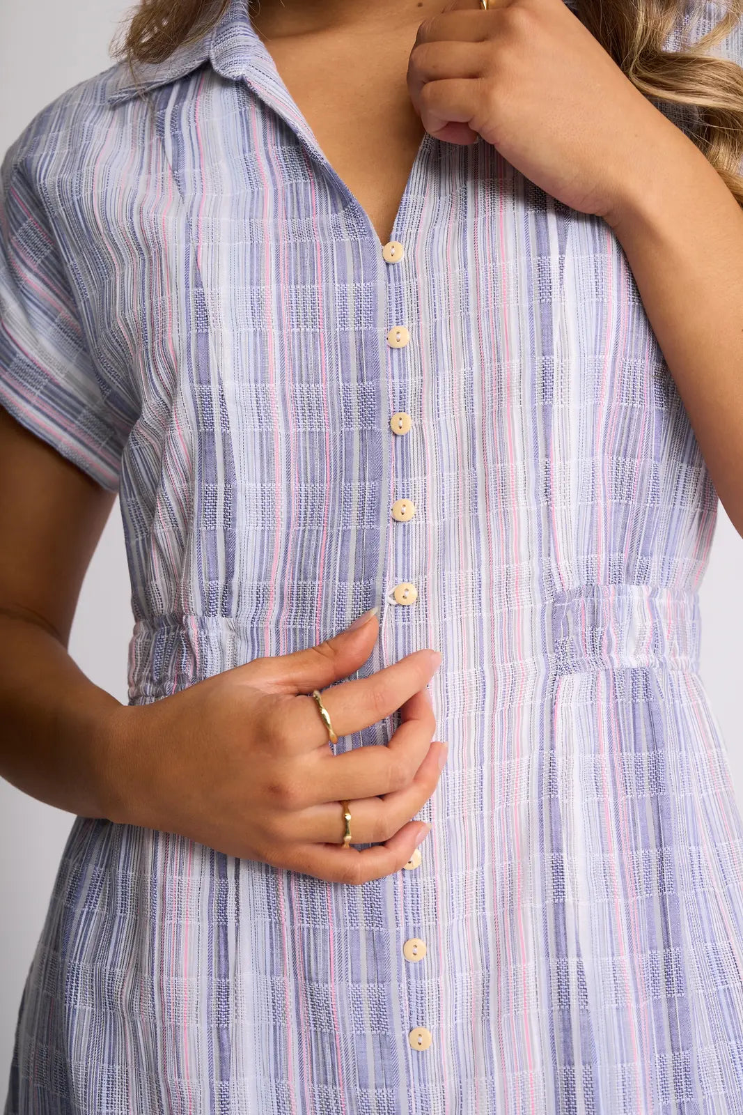 A person wearing the Vassalli Noa Dress - Blue Stripe, a short-sleeve button-down crafted from breathable cotton, holds the buttons with one hand while touching their collar with the other.