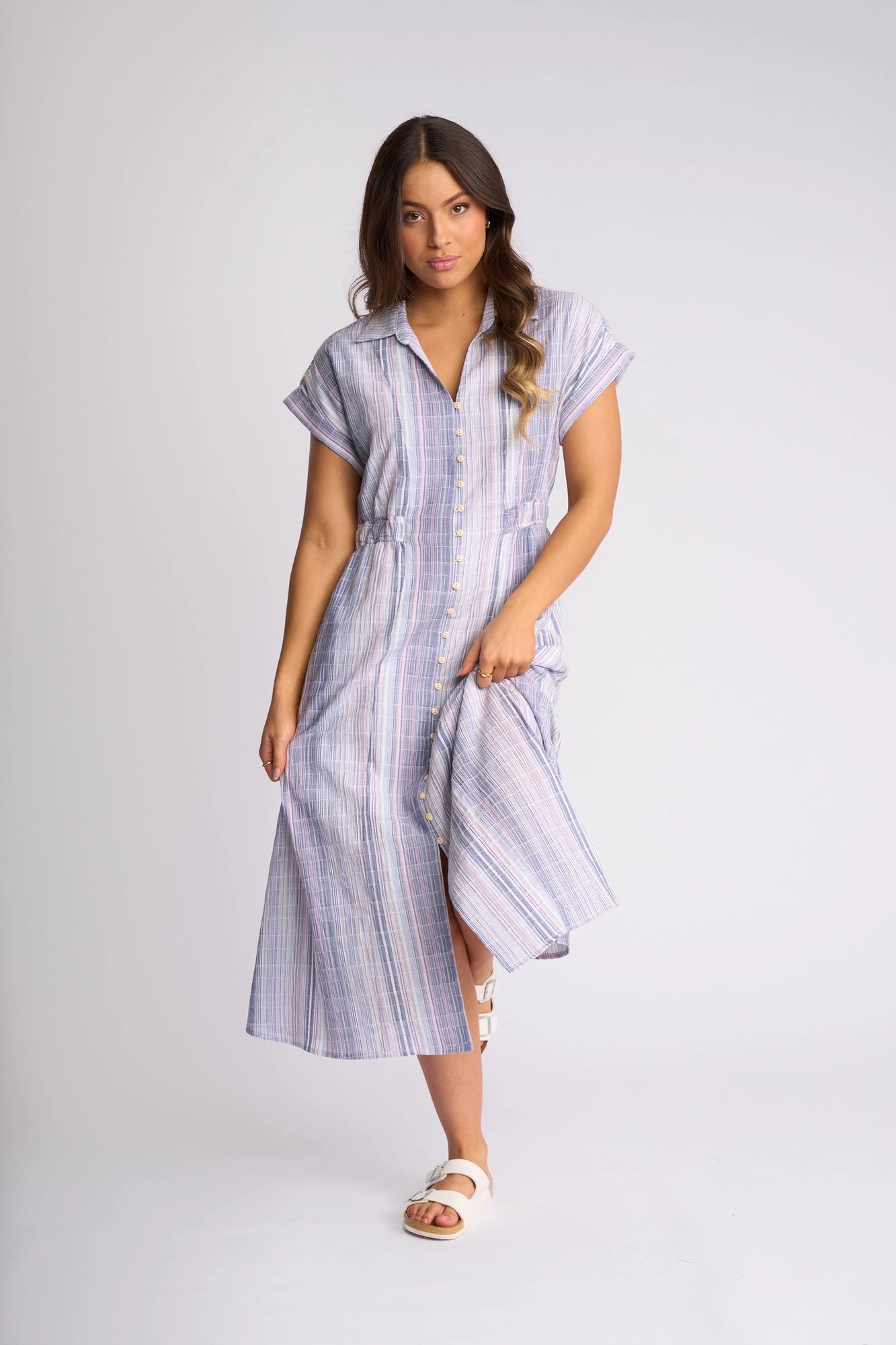 A woman wearing the Vassalli Noa Dress - Blue Stripe, a short-sleeved, button-front midi dress crafted from breathable cotton in light blue and purple stripes, paired with white sandals and standing against a plain light background.