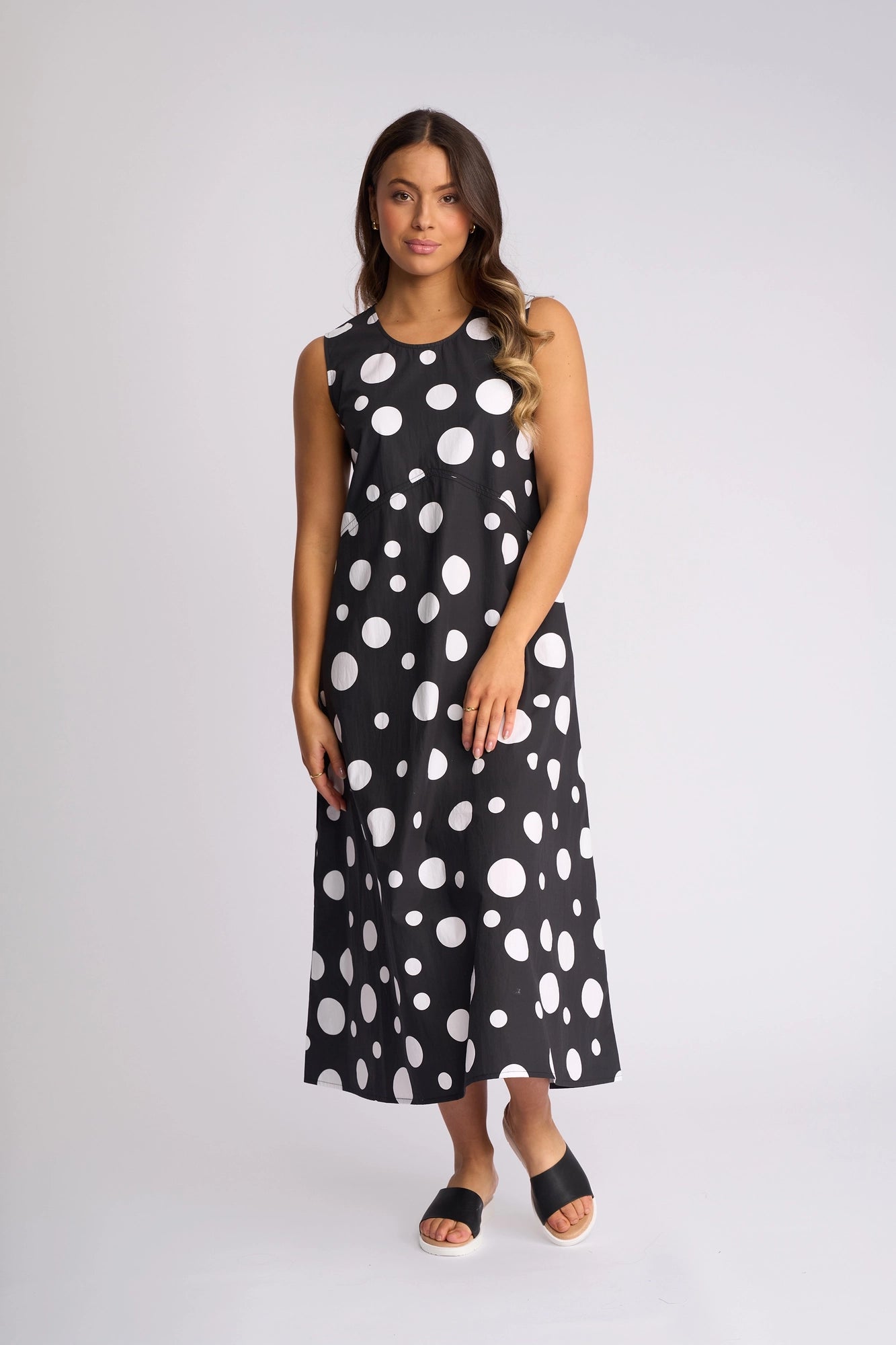 A woman wearing the Vassalli Blake Dress - Spot, a sleeveless cotton dress featuring large white polka dots, paired with black open-toe sandals, stands against a plain light background.