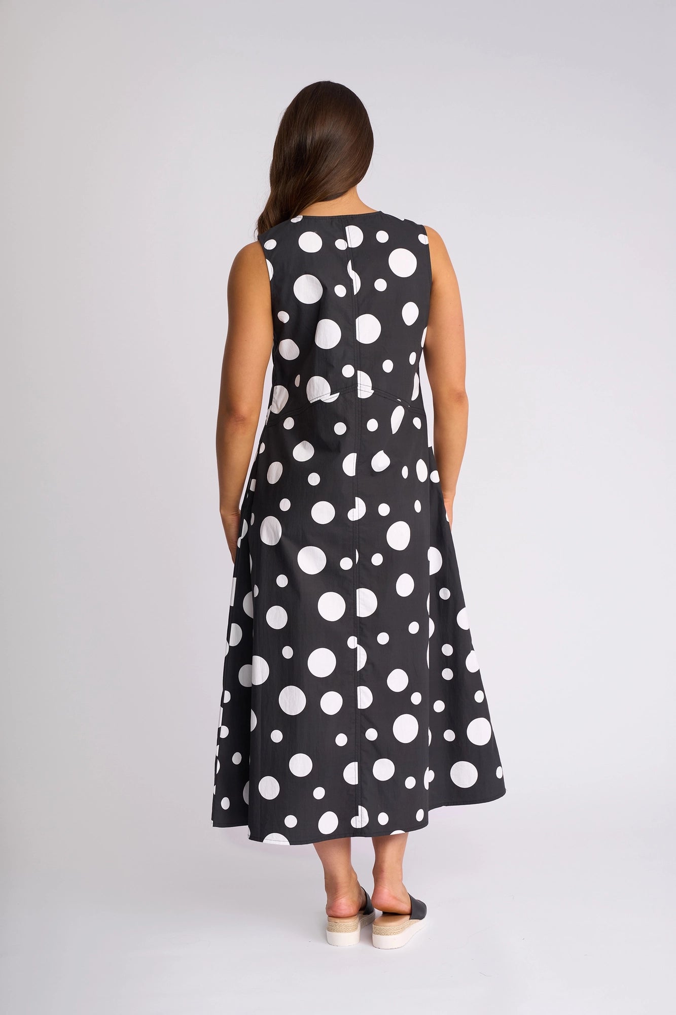 A person with long brown hair stands with their back to the camera against a plain background, wearing the Vassalli Blake Dress - Spot, a sleeveless cotton dress featuring large white polka dots on black.
