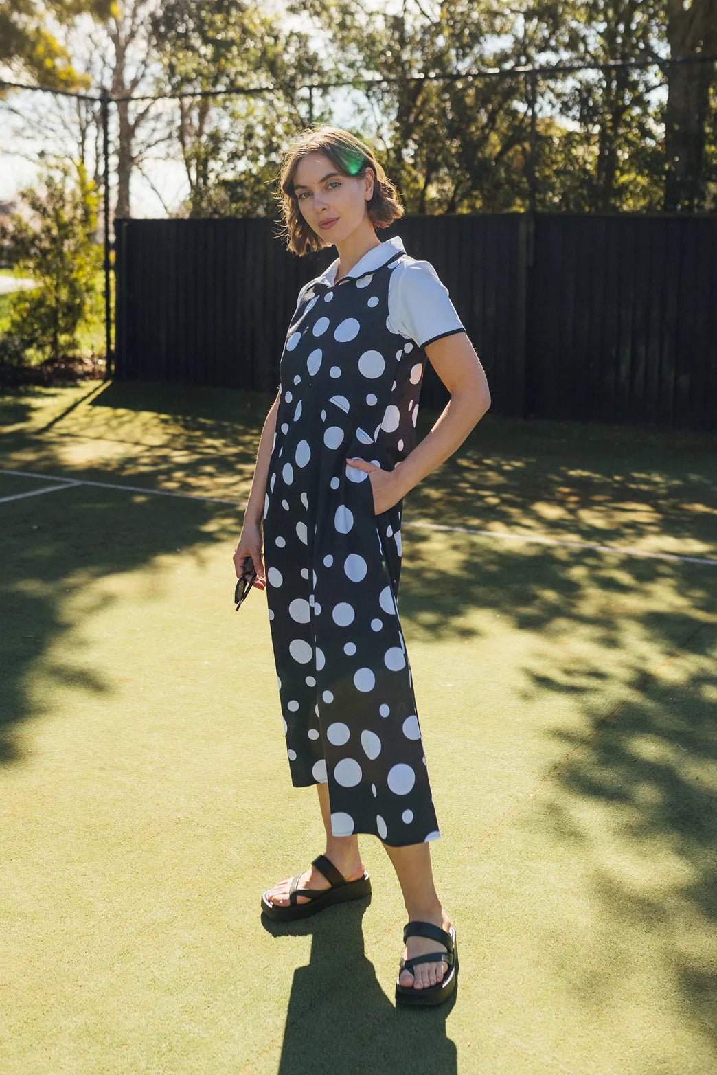 A woman stands on a sunny outdoor court, wearing the Vassalli Blake Dress in a bold spot print, layered over a white shirt and paired with sandals. She holds sunglasses, with trees and a dark fence visible in the background.