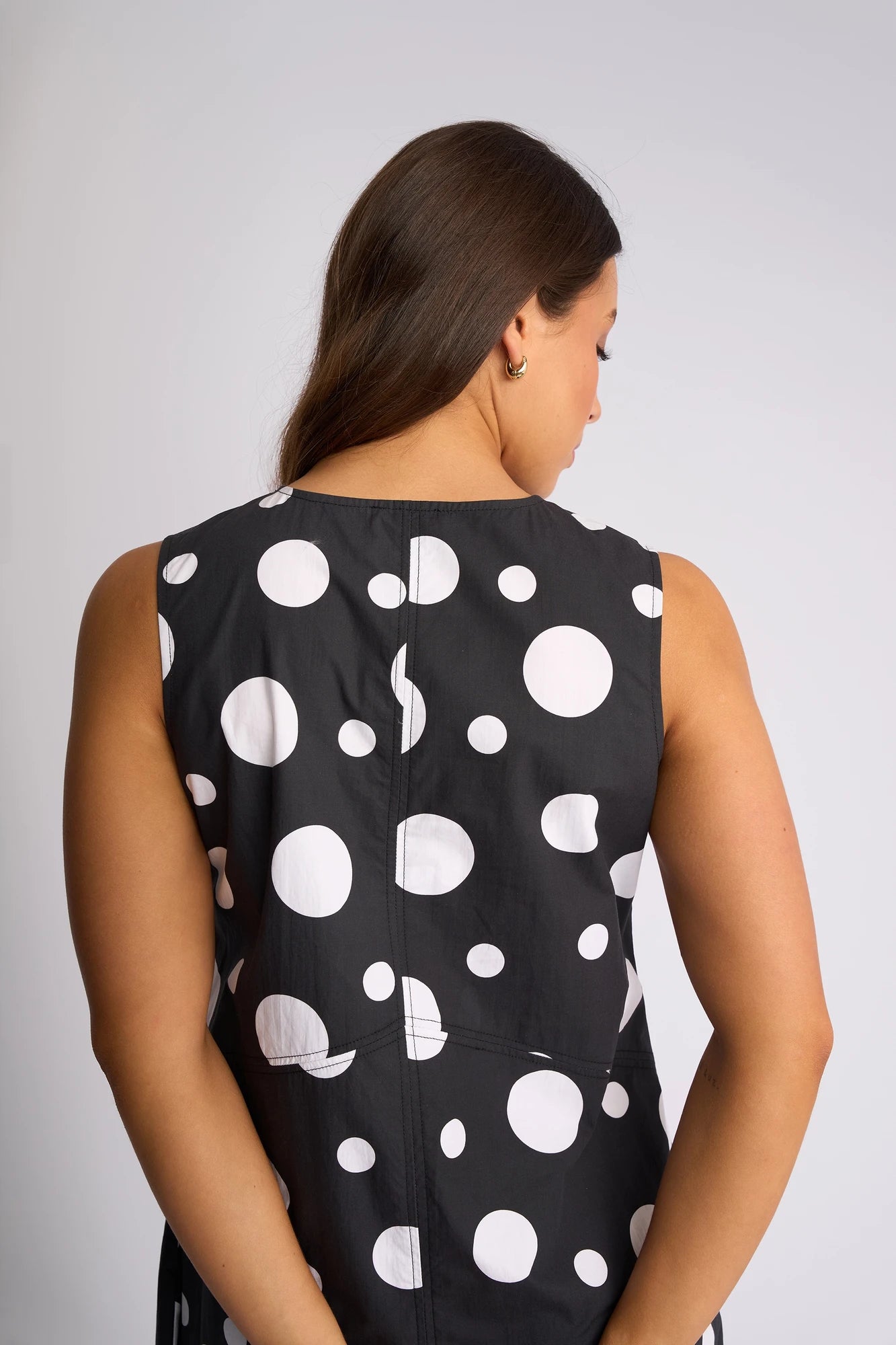 A woman with long brown hair stands facing away, wearing the Vassalli Blake Dress - Spot, a sleeveless cotton dress featuring large black and white polka dots, hands clasped behind her back, in front of a plain light background.