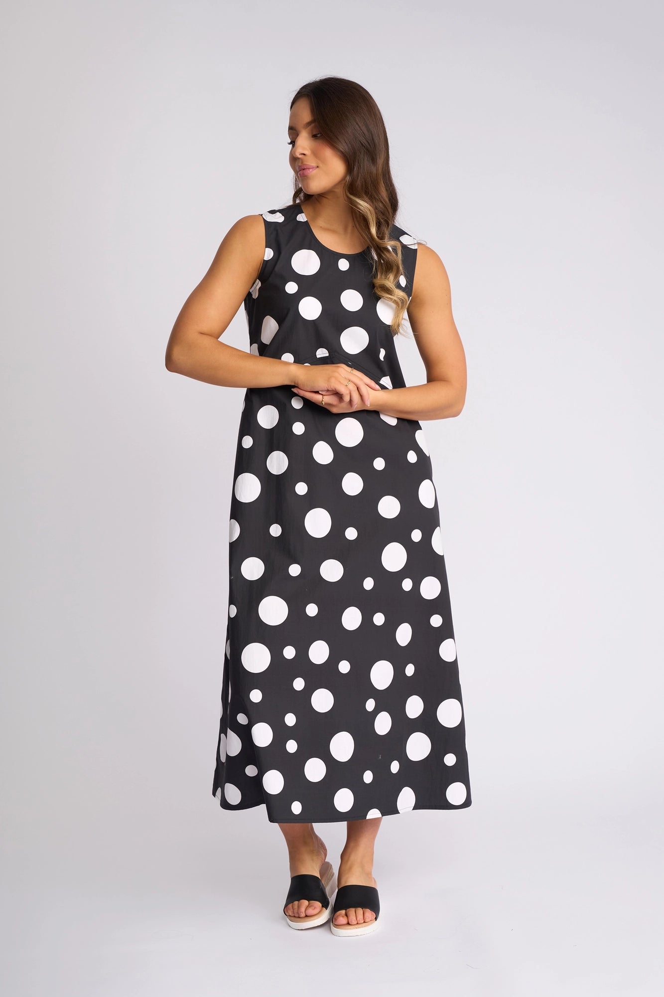 A woman wearing the Vassalli Blake Dress - Spot, an ankle-length sleeveless cotton dress with bold white polka dots on black, stands against a plain white background with her hands clasped in front, gazing to her left.