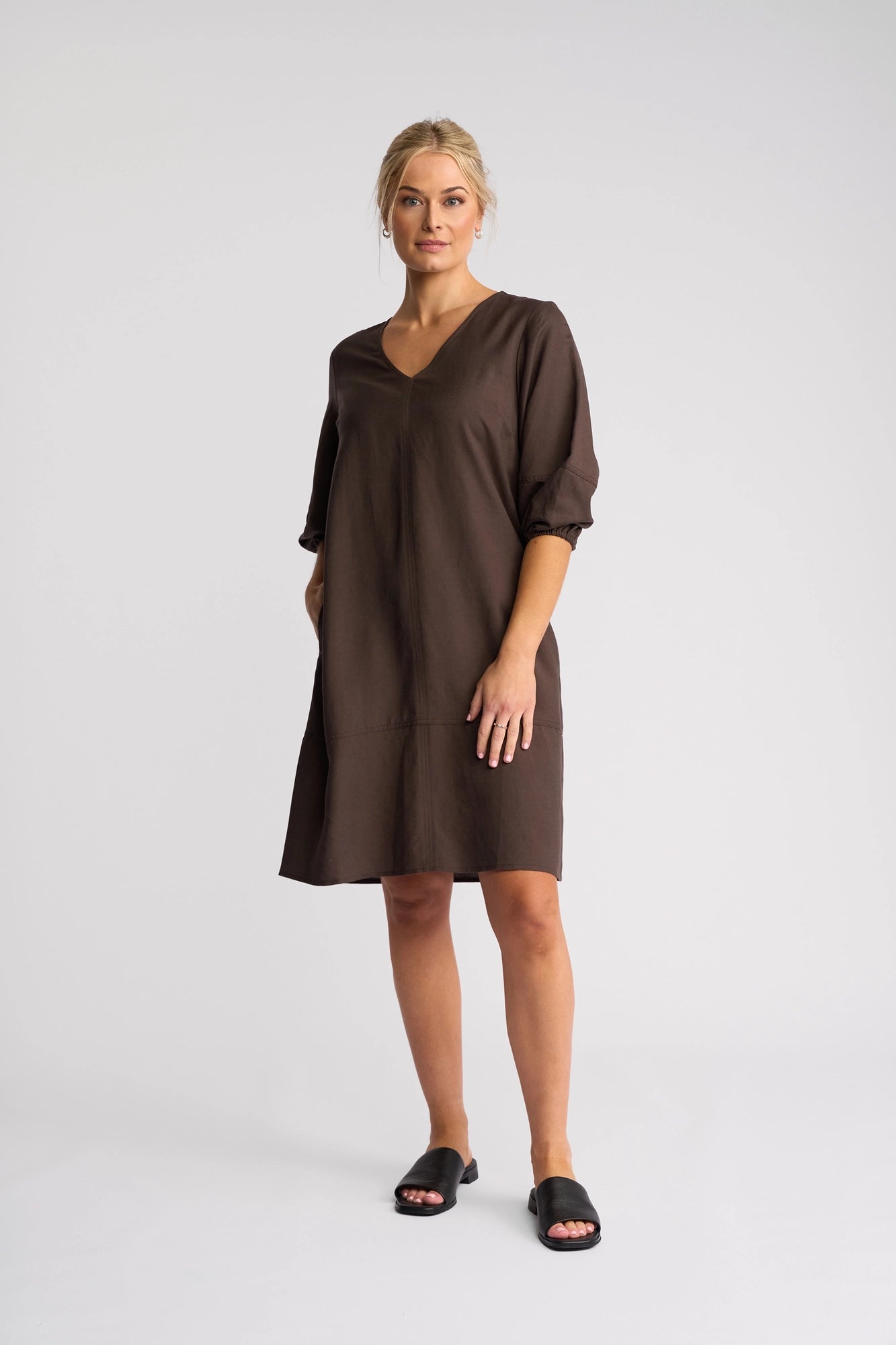 A woman stands against a plain background wearing the Vassalli Newman Dress in Chocolate—a loose, knee-length shift dress crafted from a linen-viscose blend with elbow-length sleeves—paired with black slide sandals. Her blond hair is pulled back, and she has a neutral expression.
