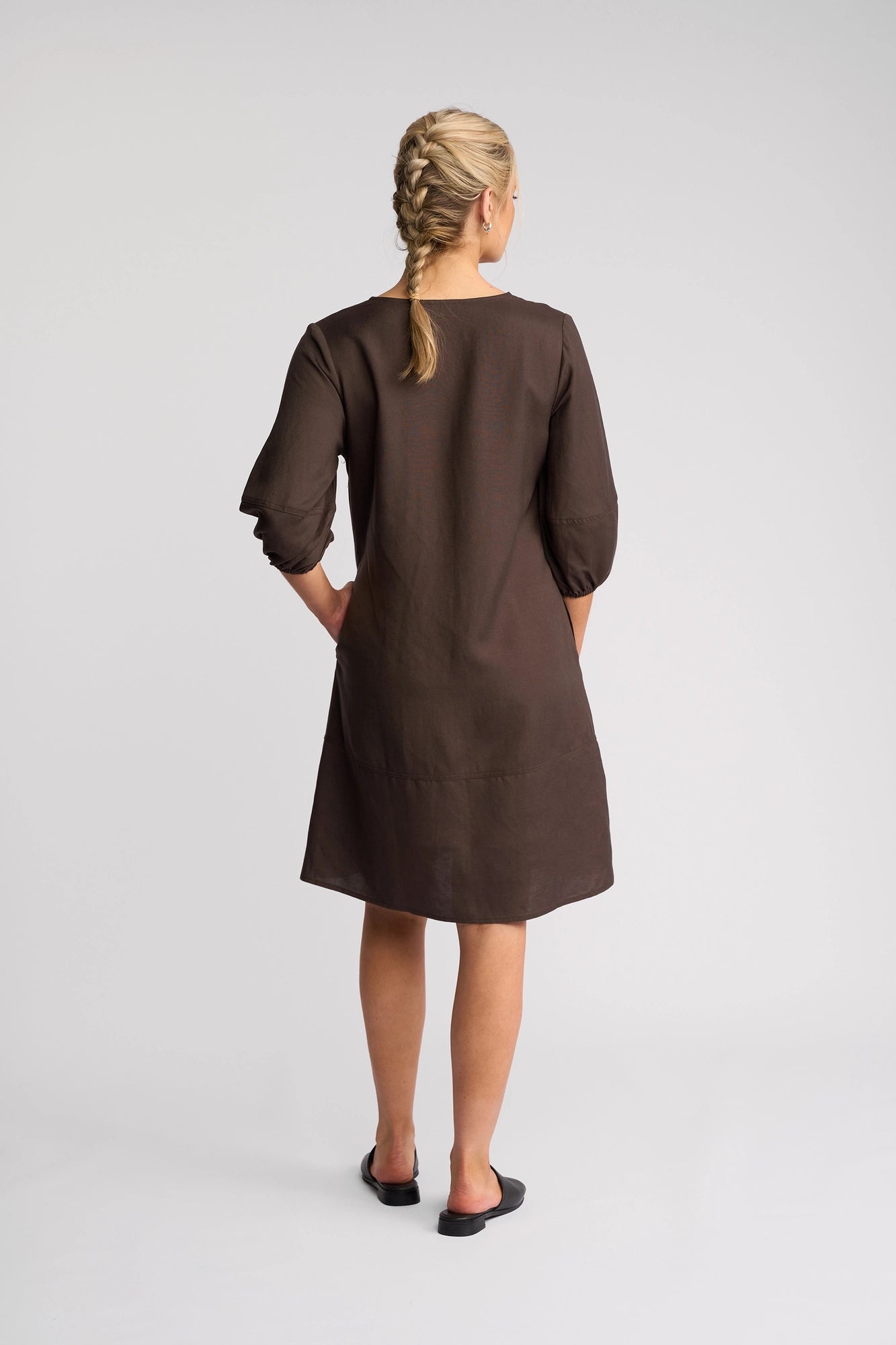 A woman with blonde hair in two braids stands with her back to the camera against a plain light background, wearing the Vassalli Newman Dress in Chocolate—a loose, knee-length linen-viscose shift dress—paired with black flats.