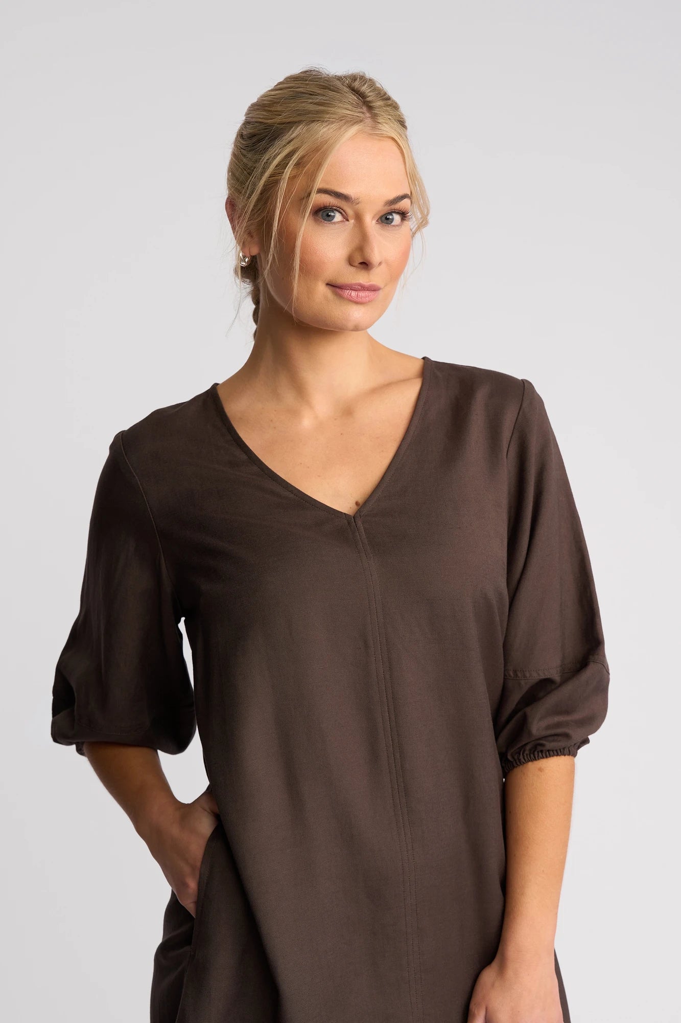 A woman with blonde hair in a low bun wears the Vassalli Newman Dress in Chocolate—a brown, V-neck linen-viscose shift dress with three-quarter sleeves—standing against a plain white background and smiling slightly.