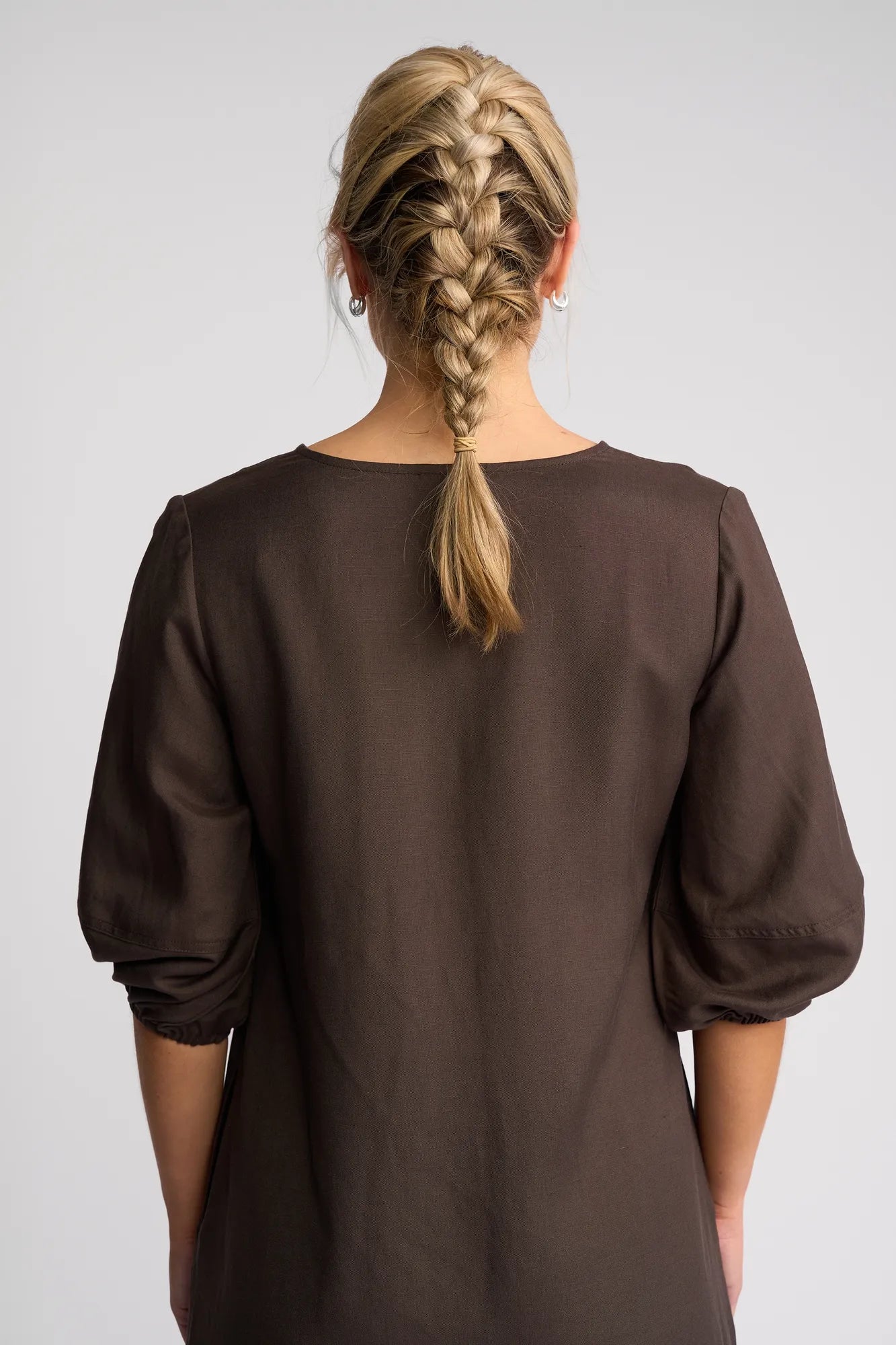 A woman with blonde hair styled in a French braid is shown from the back against a plain light background, wearing the Vassalli Newman Dress in Chocolate.