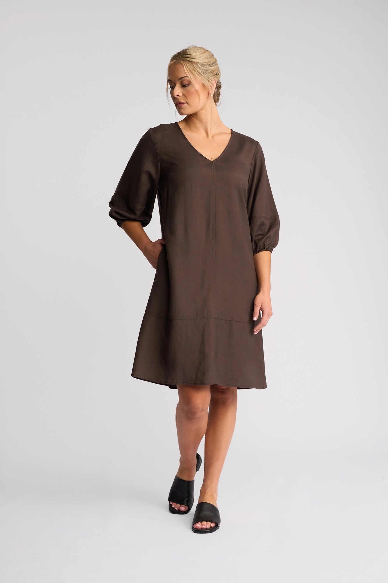 A woman stands against a plain background, looking to the side, wearing the Vassalli Newman Dress in Chocolate—a loose-fitting shift dress featuring a V-neck and three-quarter sleeves—effortlessly paired with black slide sandals.