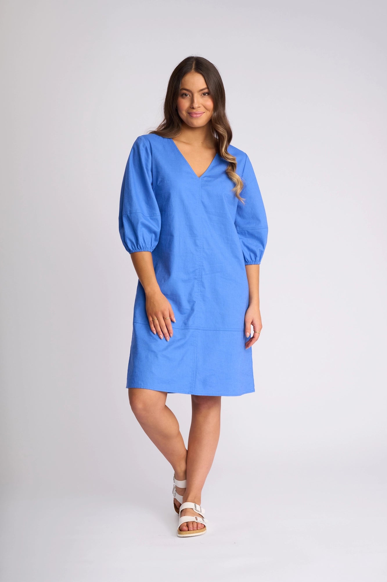 A woman stands and smiles, wearing the Vassalli Newman Dress in French Blue—a knee-length linen-viscose style with three-quarter puff sleeves—paired with white sandals and posed against a plain light background.