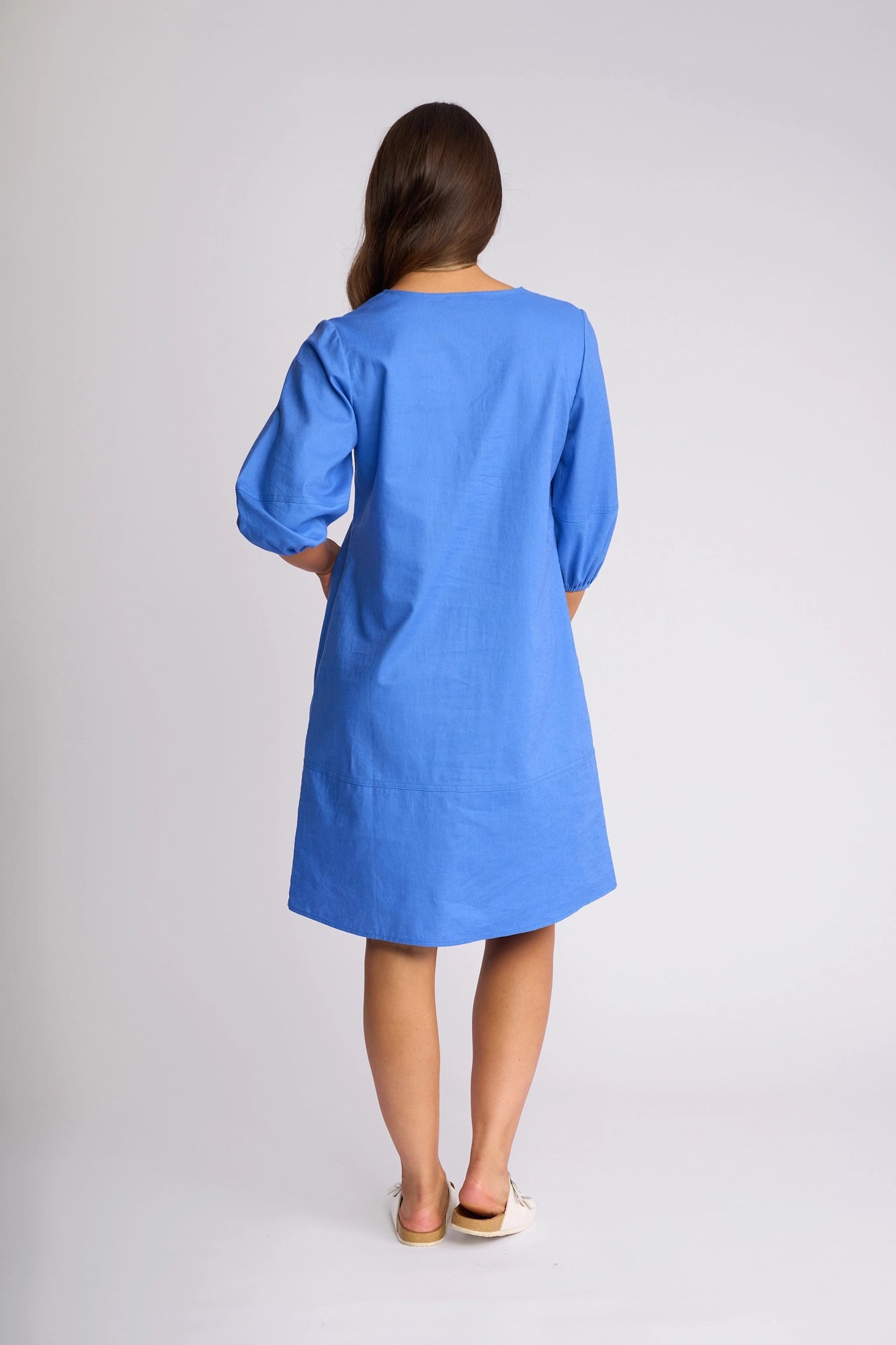 A person with long brown hair stands facing away, wearing the Vassalli Newman Dress in French Blue—a loose-fitting, knee-length shift dress with three-quarter sleeves—paired with white shoes against a plain light background.