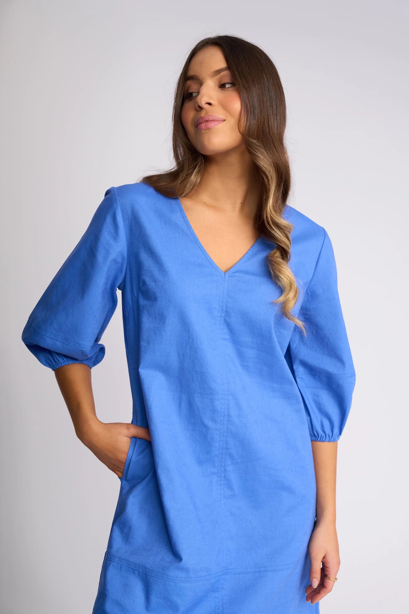 A woman wearing the Vassalli Newman Dress in French Blue—a linen-viscose shift with three-quarter sleeves—stands against a plain light background, her left hand in her pocket and head tilted slightly, long wavy hair draped over one shoulder.