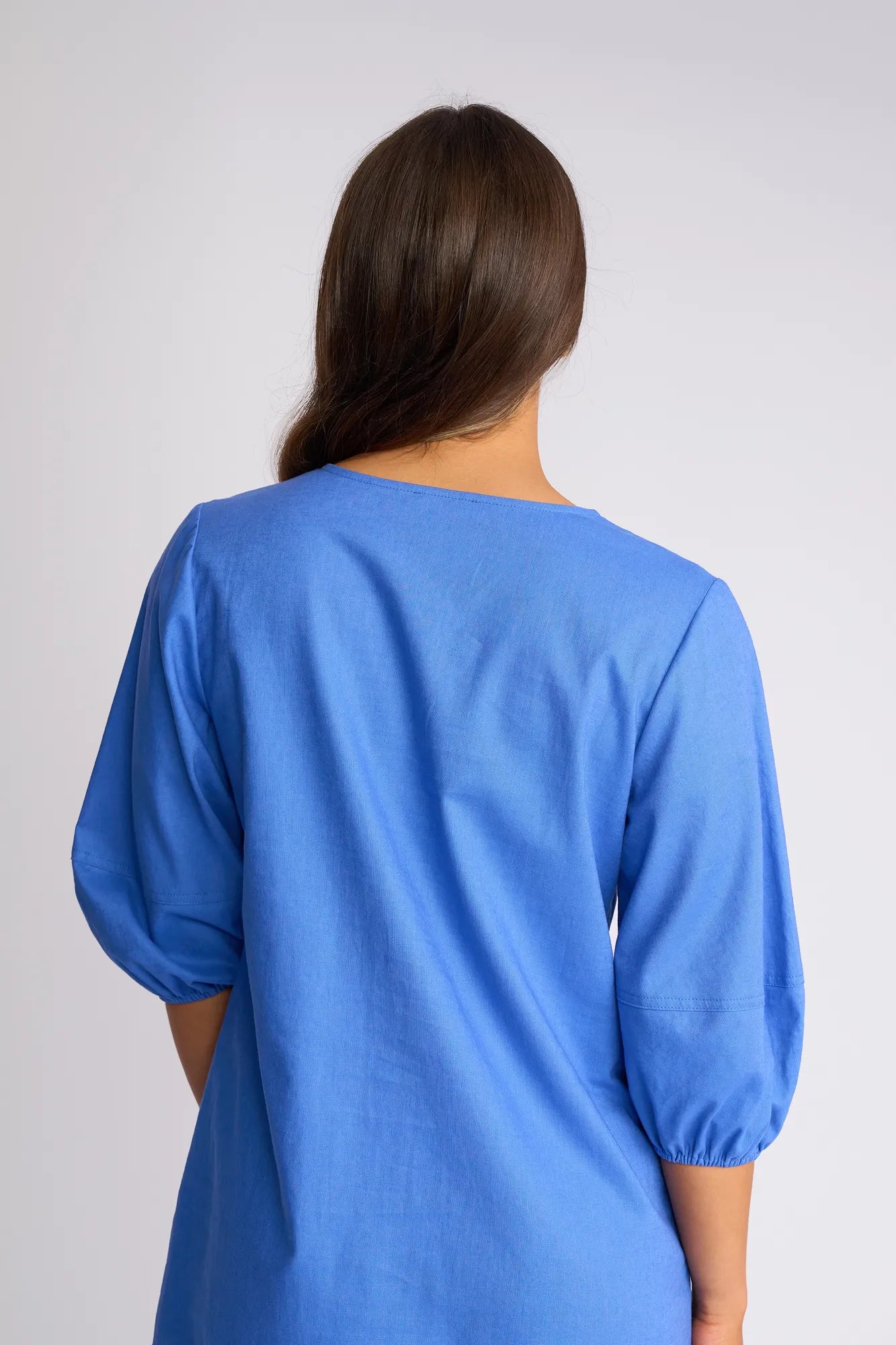 A person with long brown hair is shown from the back against a plain light background, wearing the Vassalli Newman Dress in French Blue, featuring three-quarter sleeves and a linen-viscose blend.