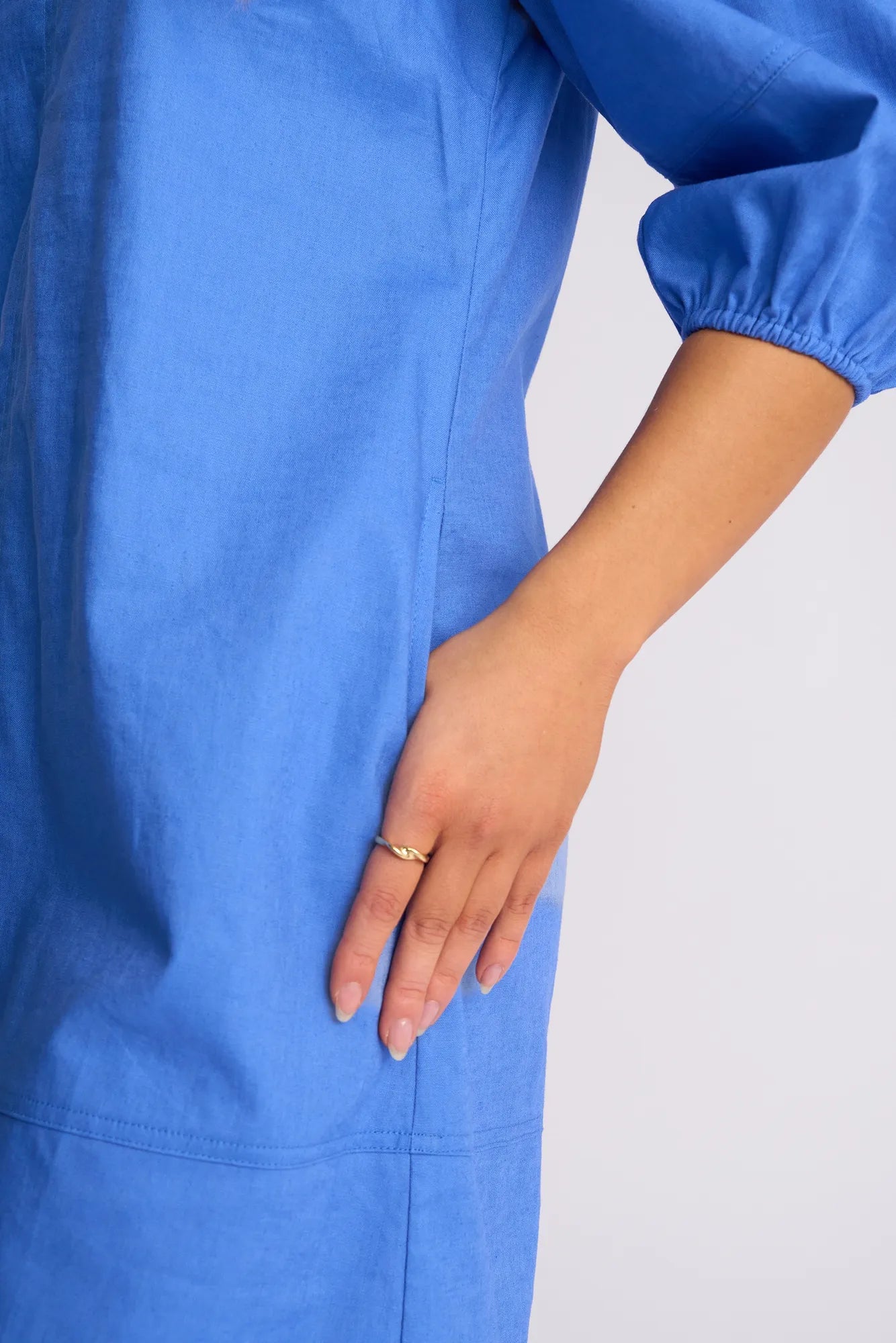 A person wearing the Vassalli Newman Dress in French Blue rests their left hand, adorned with a gold ring, on their hip against a plain, light-colored background.