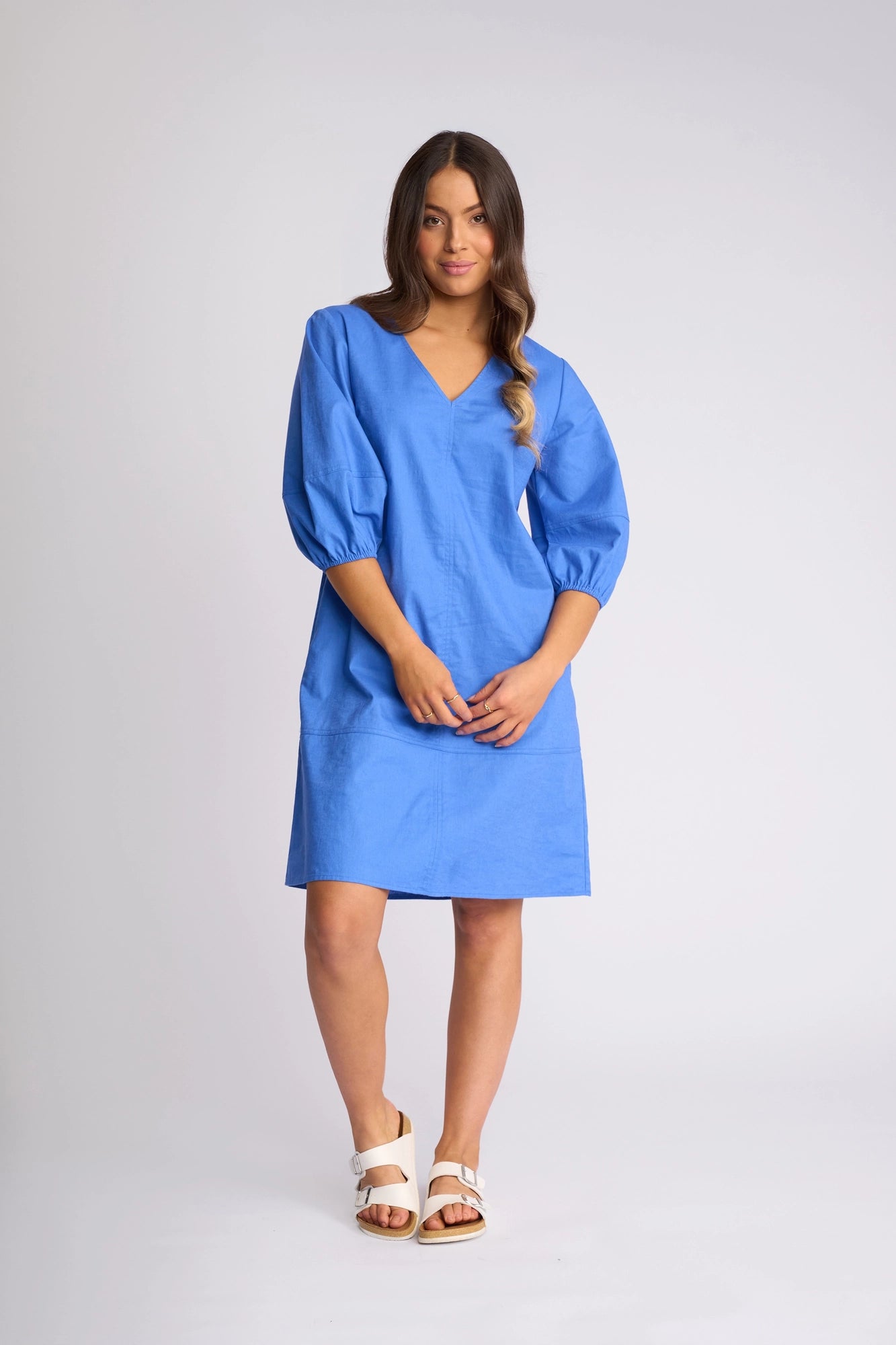 A woman stands in a studio wearing the Vassalli Newman Dress in French Blue, a knee-length shift dress featuring three-quarter puff sleeves. She pairs it with white sandals. The background is plain and light-colored.