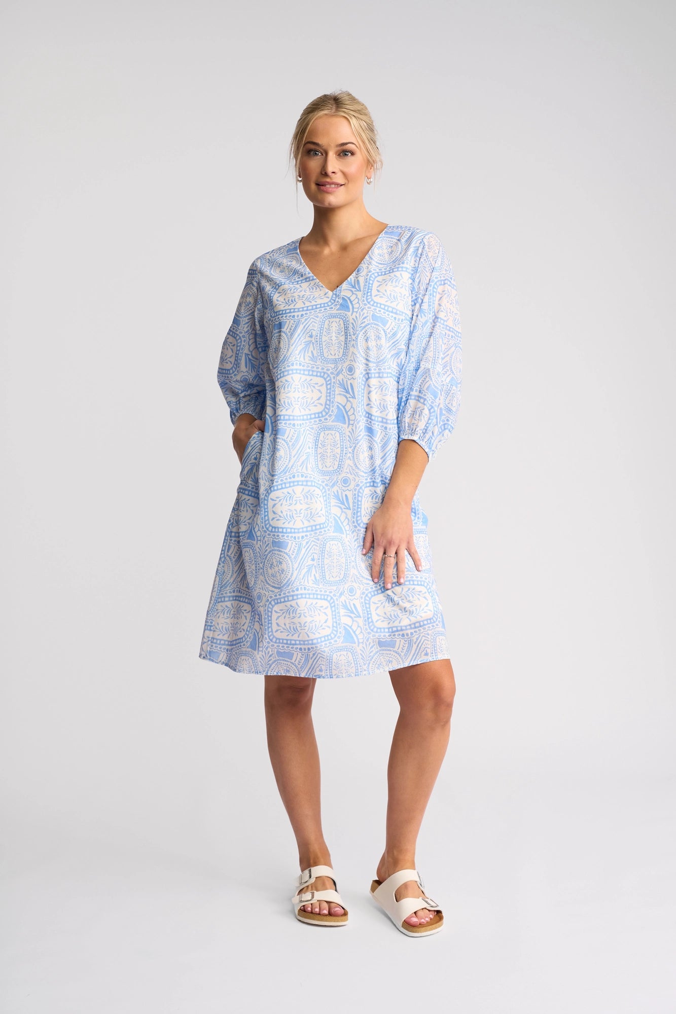 A woman stands against a plain background wearing the Vassalli Newman Dress - Palma, which features a Mediterranean tile print in light blue, three-quarter sleeves, and pockets. Made from 100% cotton, the dress is perfectly paired with white sandals. Her blonde hair is tied back as she looks forward with a slight smile.