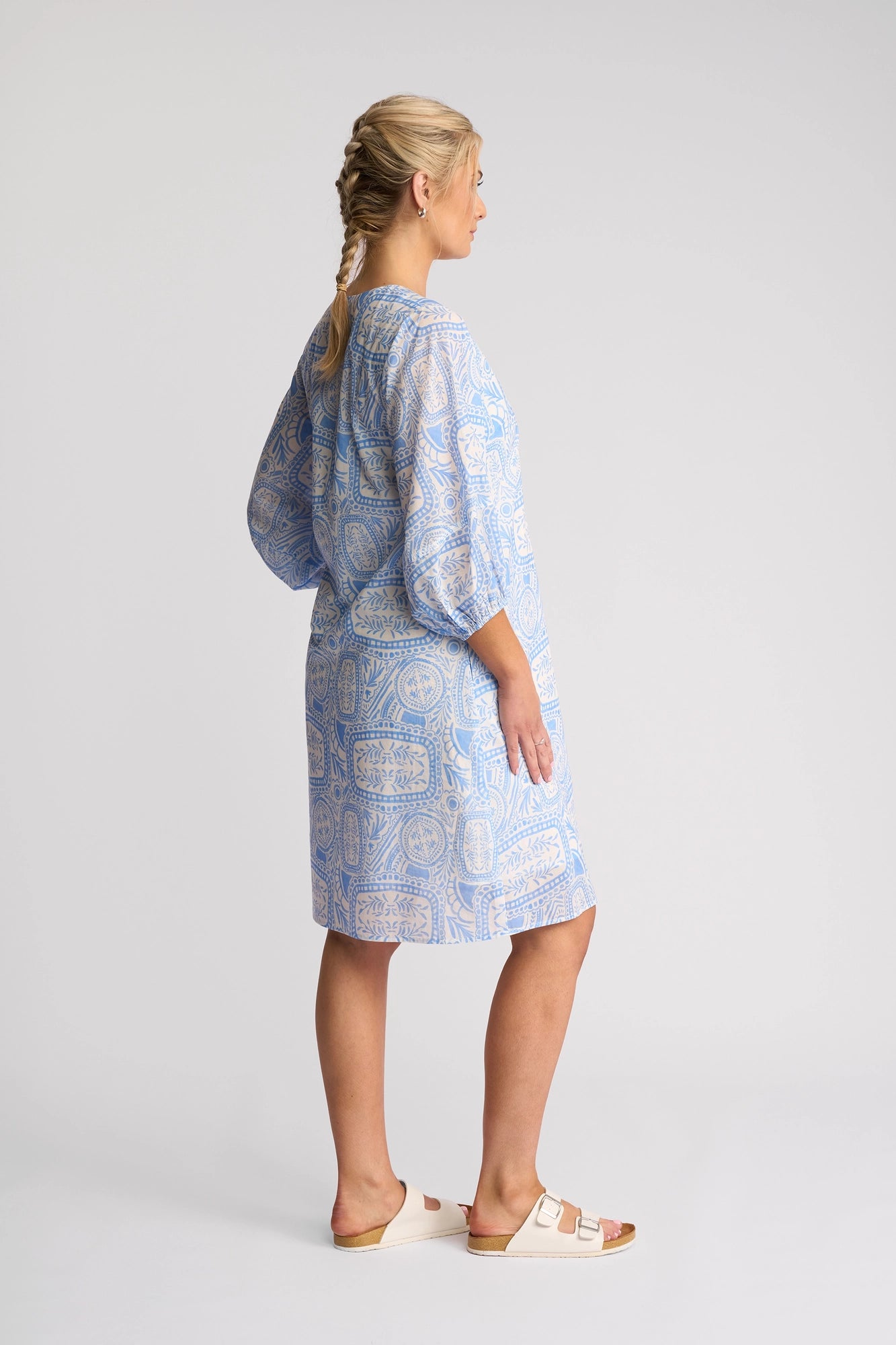 A woman with braided blonde hair wears a loose, light blue Vassalli Newman Dress - Palma featuring a Mediterranean tile print, crafted from 100% cotton. She pairs it with white sandals and stands sideways against a plain light background.