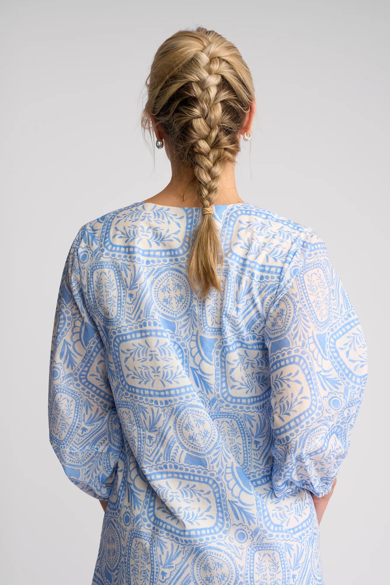 A woman with blonde hair in a French braid is shown from the back against a plain background, wearing the Vassalli Newman Dress - Palma, made from 100% cotton and featuring a light blue and white Mediterranean tile print.