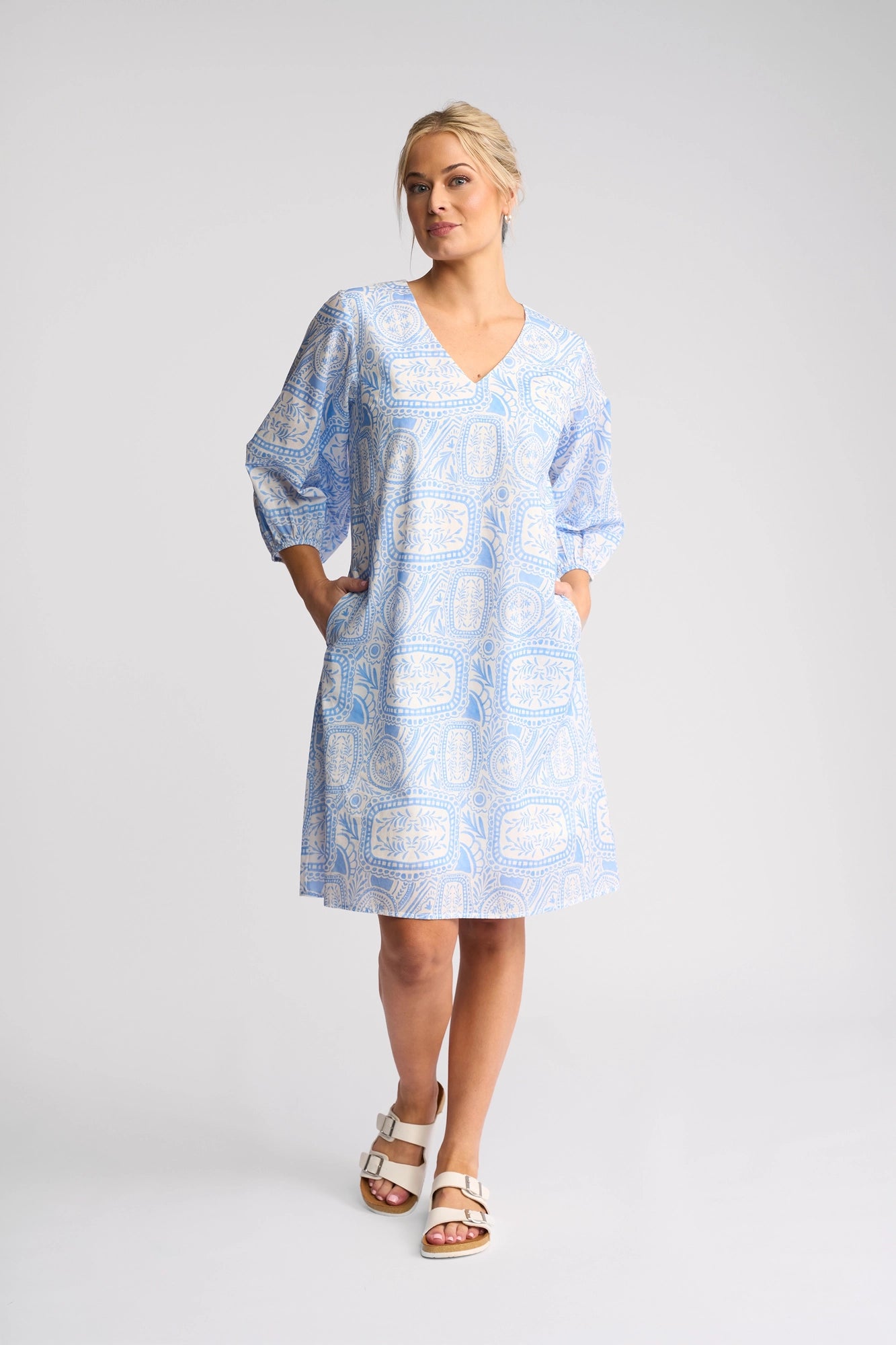 A woman stands against a plain background wearing the Vassalli Newman Dress - Palma, a loose, light blue dress featuring a beautiful Mediterranean tile print. Made from 100% cotton, the dress has elbow-length sleeves and is styled with white sandals. She has blonde hair in a low bun and her hands are tucked into the dress pockets.