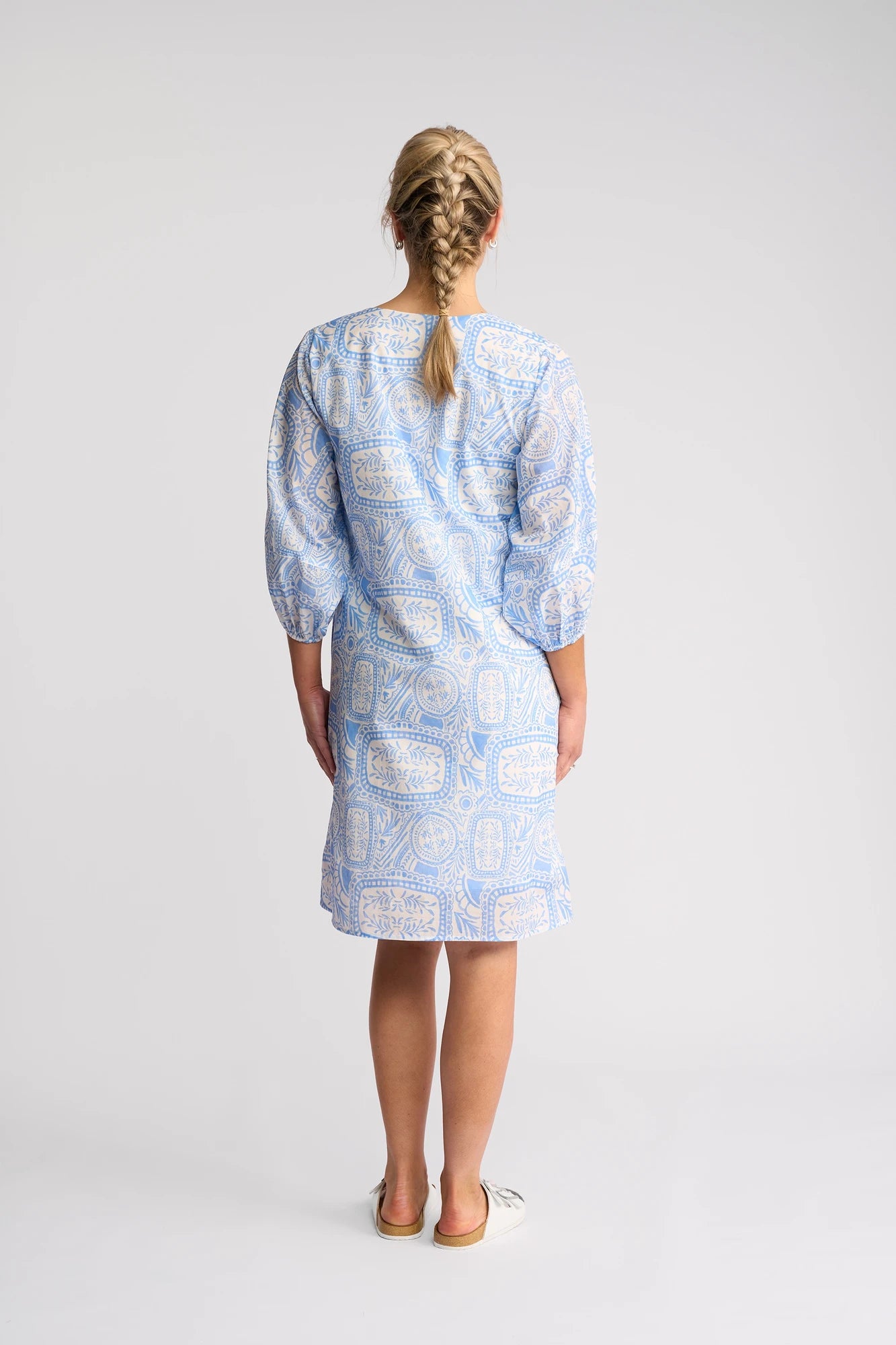 A woman with blonde braided hair wears the Vassalli Newman Dress - Palma, showcasing a striking Mediterranean tile print in blue and white on 100% cotton fabric, as she stands facing away from the camera against a plain white background.