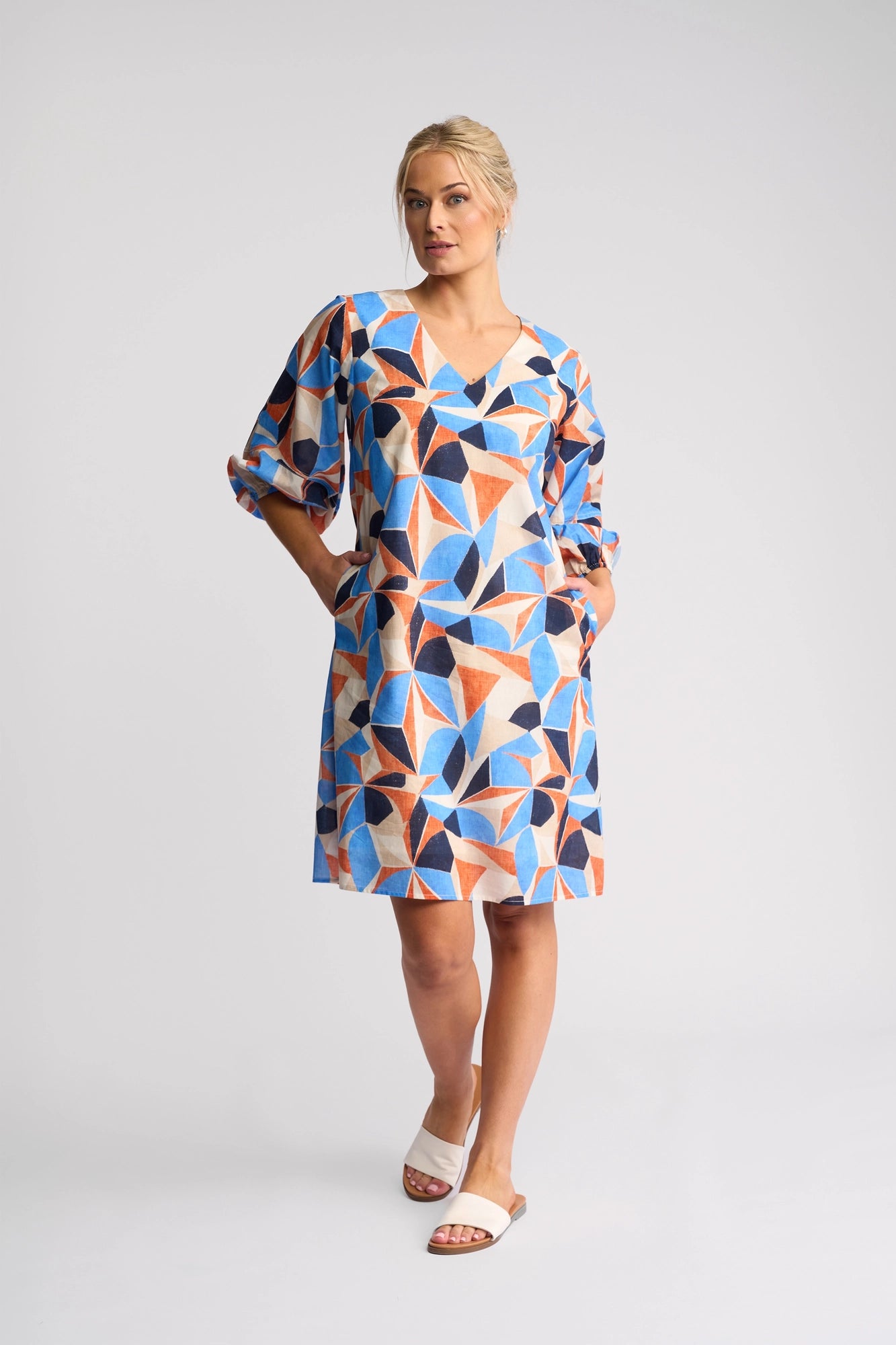A woman stands with her hands in the pockets of a Vassalli Newman Dress - Row, a 100% cotton geometric print dress adorned with bold blue, orange, and beige shapes. She looks to the side and pairs the dress with white slide sandals against a plain, light-colored background.