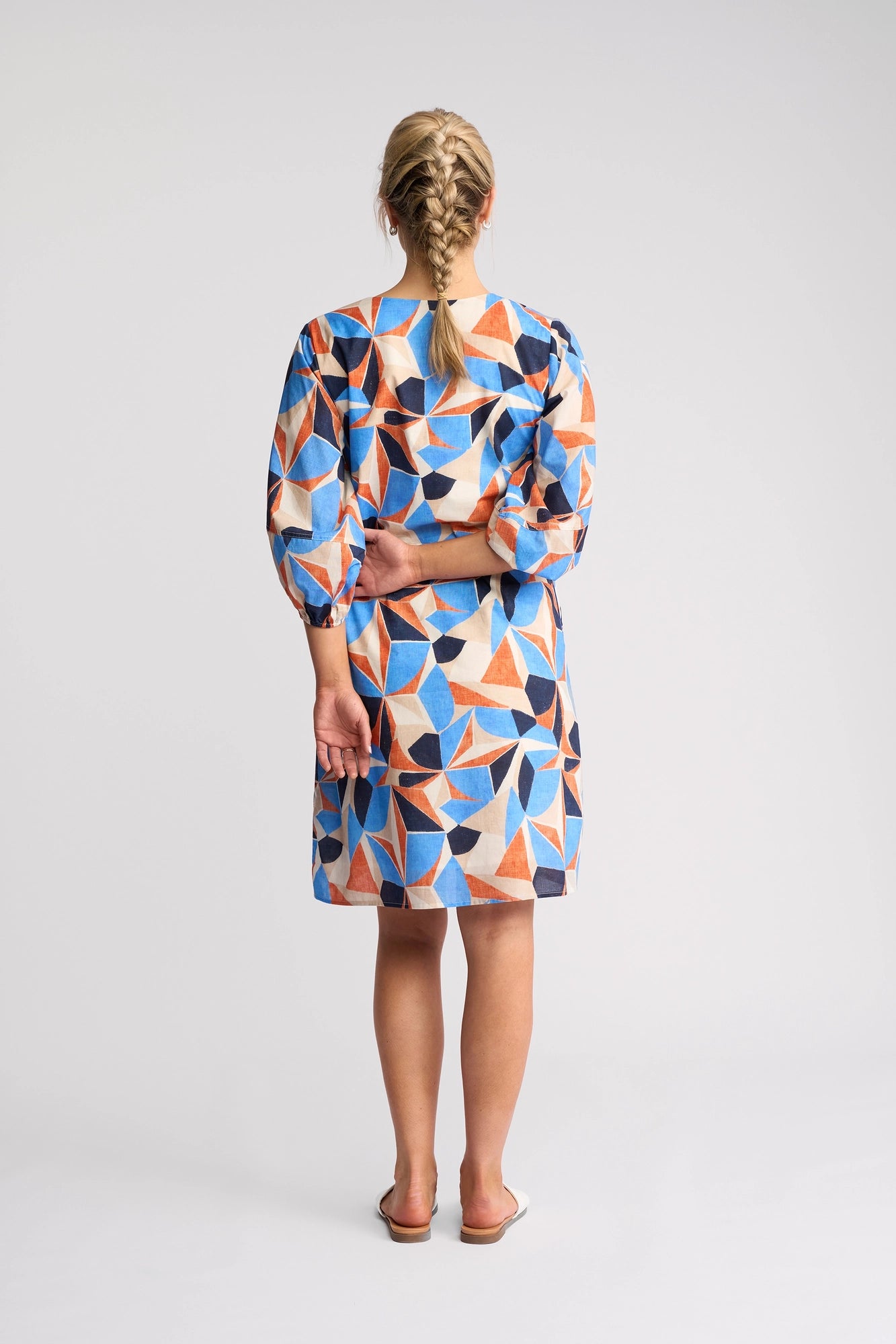 A woman with blond braided hair stands barefoot, facing away, wearing the Vassalli Newman Dress - Row. This knee-length dress features a bold geometric blue, orange, and beige pattern that flatters her silhouette. Made from 100% cotton, the stylish geometric print dress enhances her figure as she holds her left arm behind her back against a plain background.
