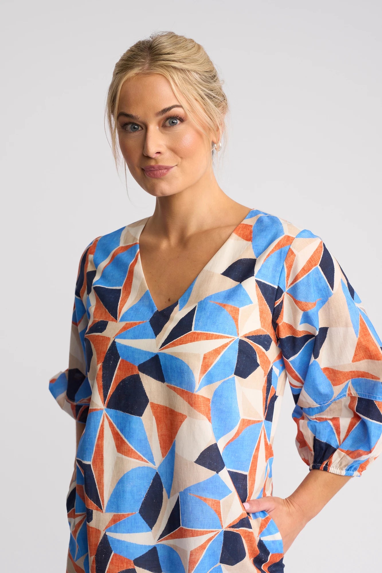 A woman with blonde hair in a low bun wears the Vassalli Newman Dress - Row—a 100% cotton geometric print dress in blue, orange, and beige—as she stands against a plain light background with one hand in her pocket.