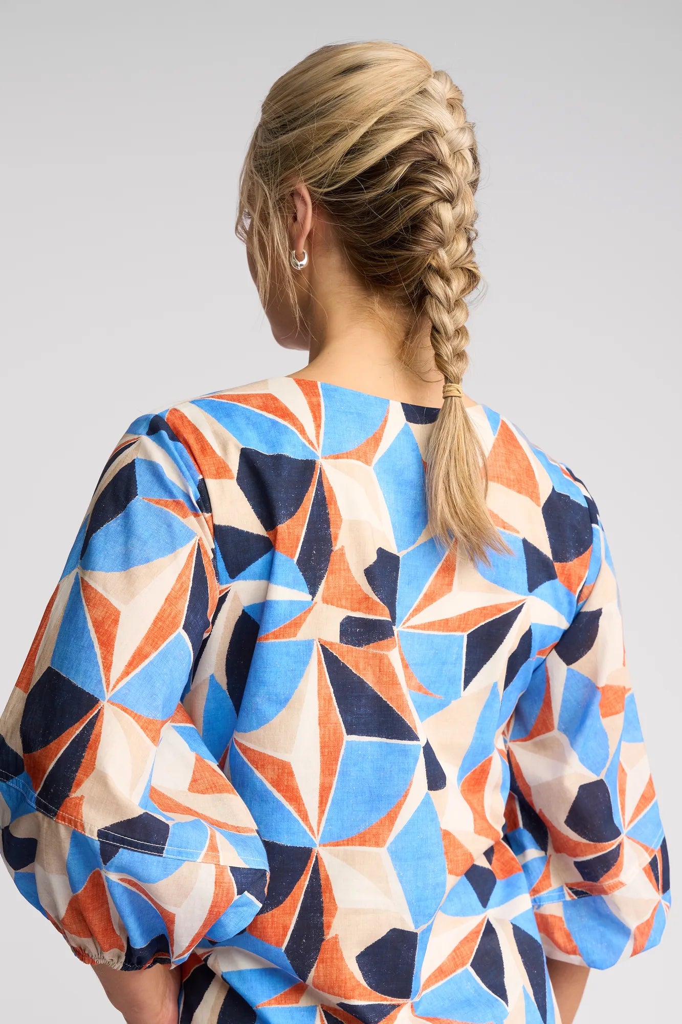 A person with blonde hair styled in a braid is shown from the back against a simple light gray background, wearing the Vassalli Newman Dress - Row—a vibrant geometric print dress in shades of blue, orange, black, and beige crafted from 100% cotton.
