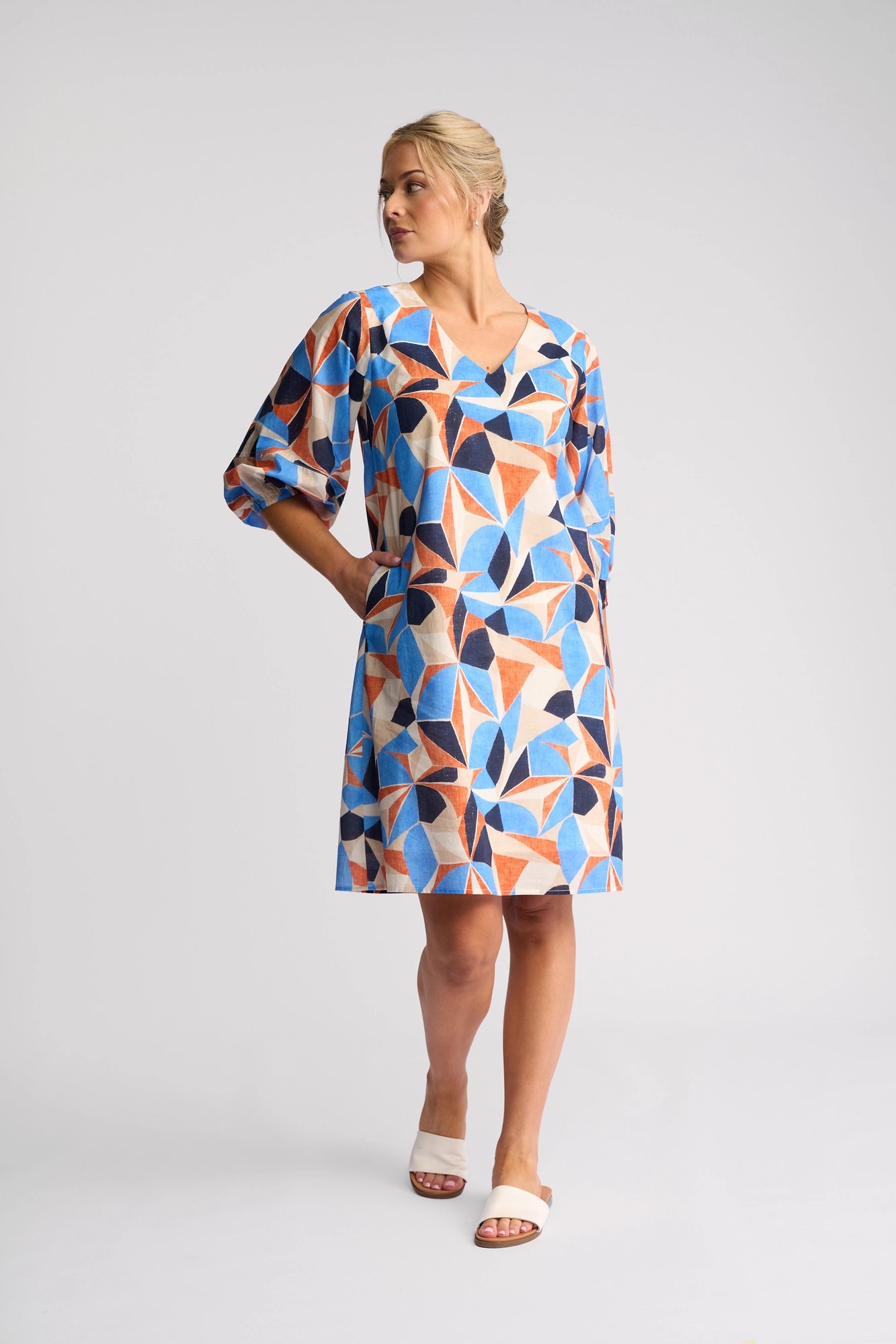 A woman stands sideways in a knee-length Vassalli Newman Dress - Row, featuring blue, orange, and beige geometric patterns. With one hand in her pocket and looking to her left, she completes the look with white slide sandals. This dress is made from 100% cotton.