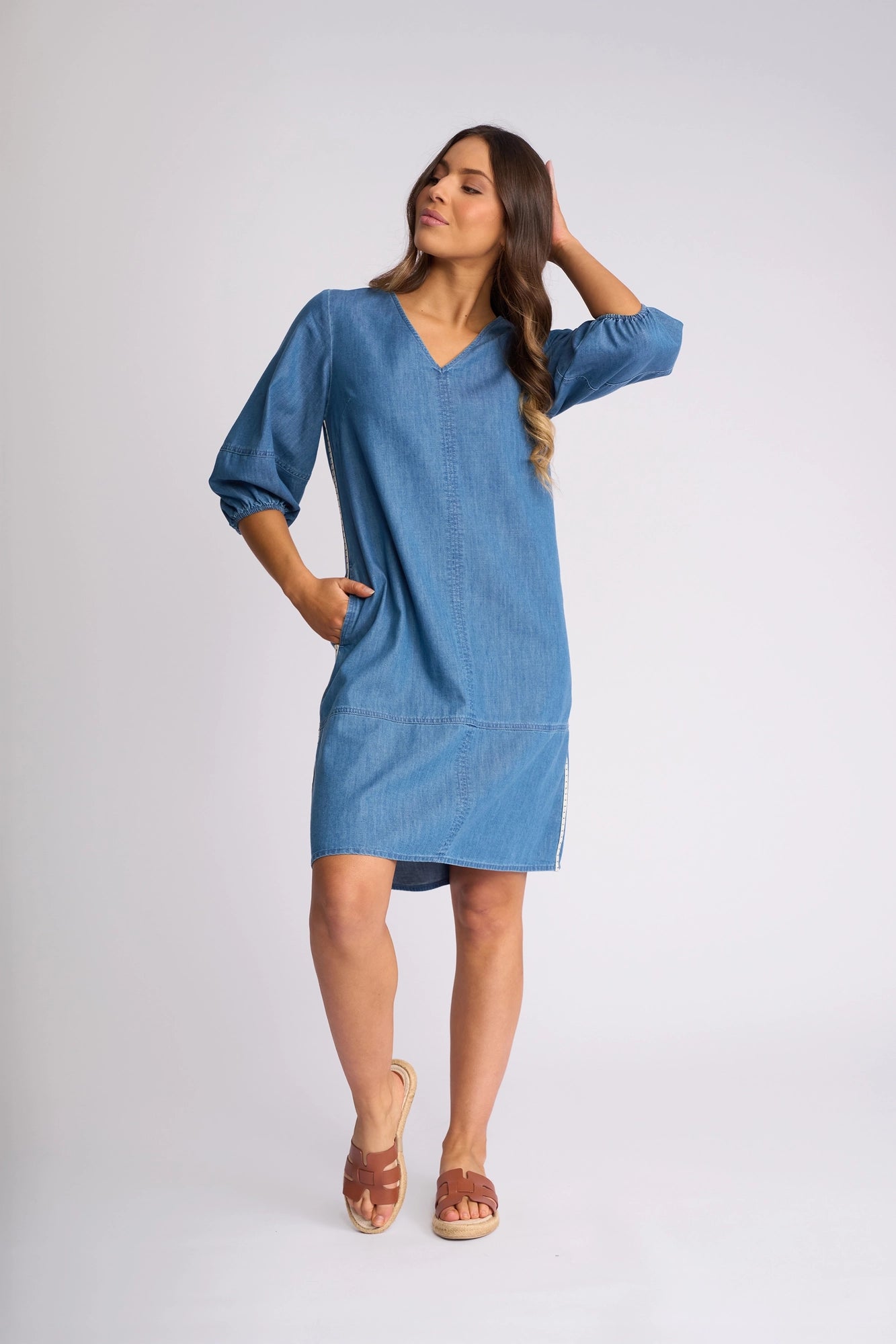 Wearing the Vassalli Nevada Dress in Surf Blue—a loose-fitting, knee-length cotton denim style with three-quarter sleeves—she stands against a plain light background, one hand in her pocket and the other brushing her hair, paired with brown sandals for an effortlessly chic summer look.