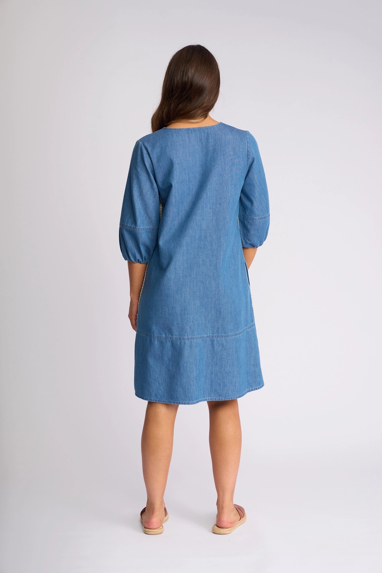 A woman stands facing away against a plain light background, wearing the Vassalli Nevada Dress in Surf Blue—a knee-length cotton denim summer dress with three-quarter sleeves—paired with beige sandals.