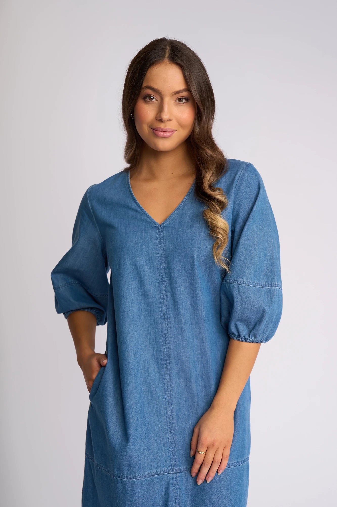 A woman with long wavy hair, wearing the Vassalli Nevada Dress in Surf Blue made from soft cotton denim with three-quarter sleeves, stands against a plain light background, smiling softly with one hand in her pocket.
