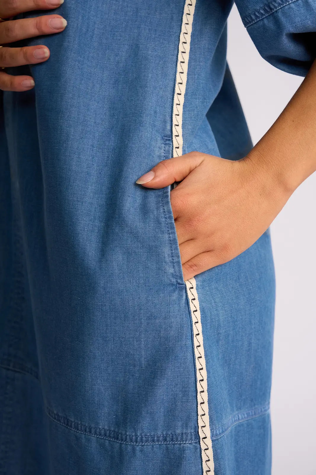 A person’s hand is partially in the pocket of the Vassalli Nevada Dress in Surf Blue, which features a white zigzag-stitched trim along the pocket seam.