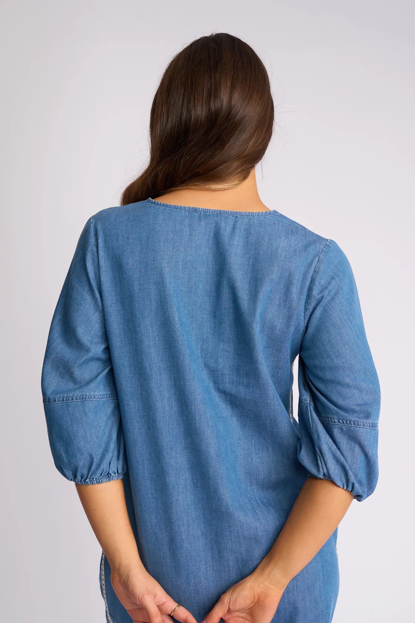 A person with long brown hair stands facing away from the camera, wearing the Vassalli Nevada Dress in Surf Blue, featuring three-quarter sleeves and cotton denim fabric, with their hands held behind their back.