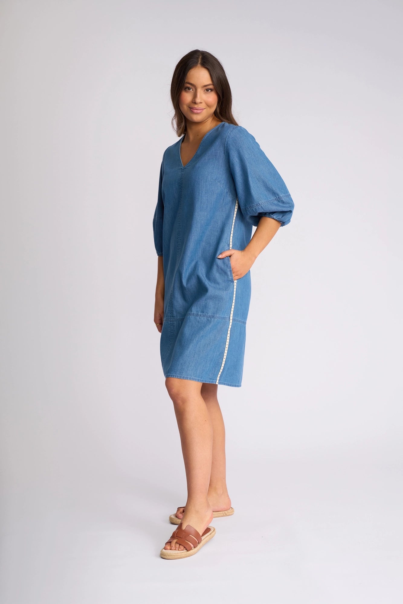 A woman stands against a plain, light background wearing the Vassalli Nevada Dress - Surf Blue—a loose-fitting cotton denim dress featuring short puff sleeves and convenient side pockets. She pairs it with brown sandals and offers a subtle smile, showcasing an effortlessly stylish look ideal for summer.