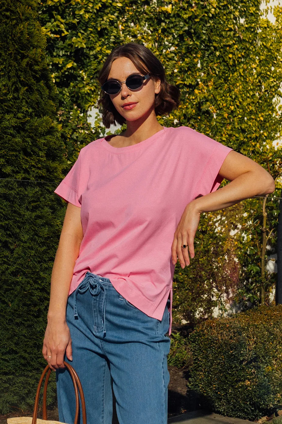A woman wearing sunglasses stands outdoors in sunlight, holding a brown handbag. She is dressed in the Vassalli Thea Top - Sweet Pink, which features a relaxed fit and pairs effortlessly with blue jeans. Green foliage and hedges serve as the backdrop.