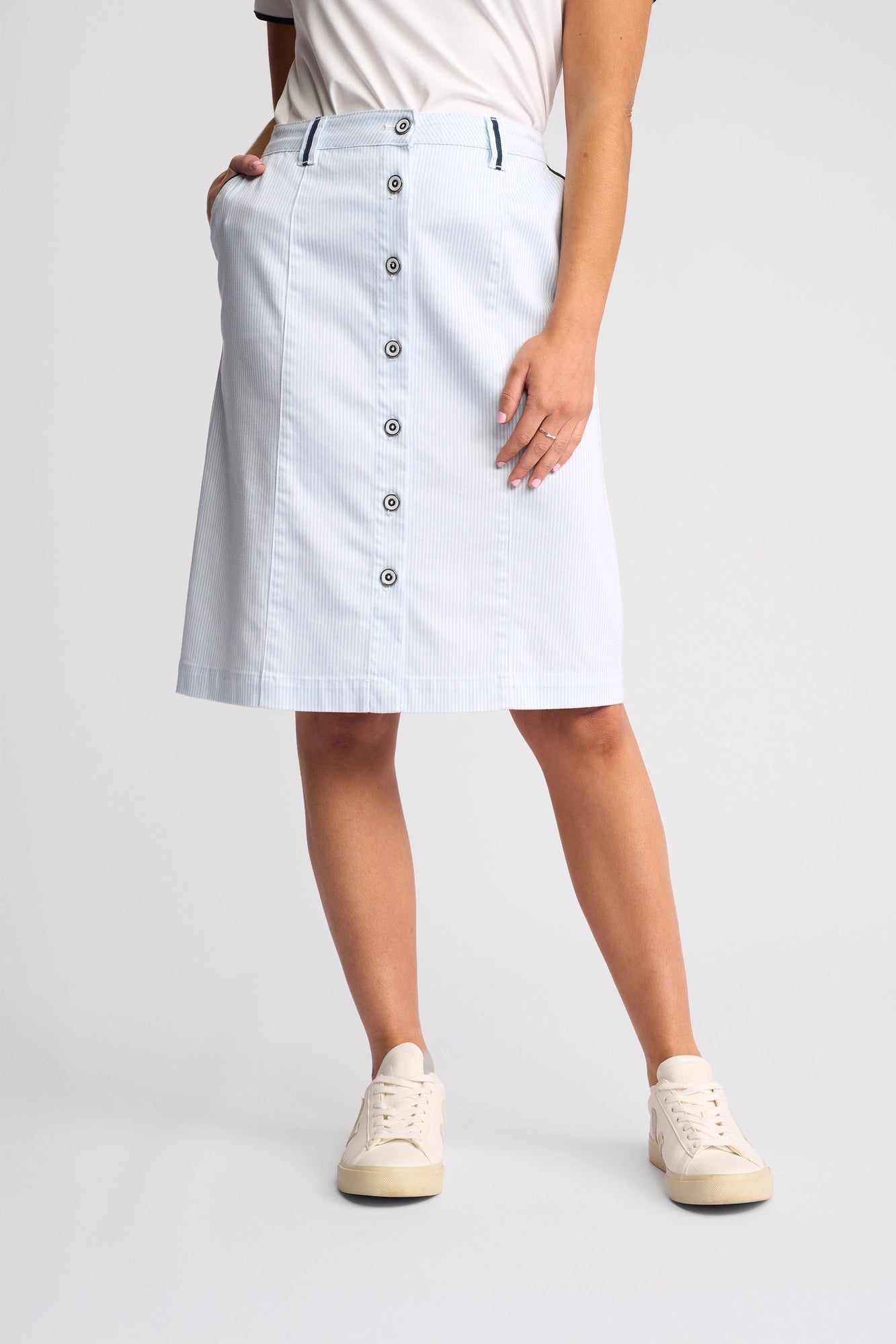 A person wearing the Vassalli Orion Skirt in Blue Stripe Denim, styled with a white t-shirt and cream sneakers, stands with one hand in a pocket against a plain background.