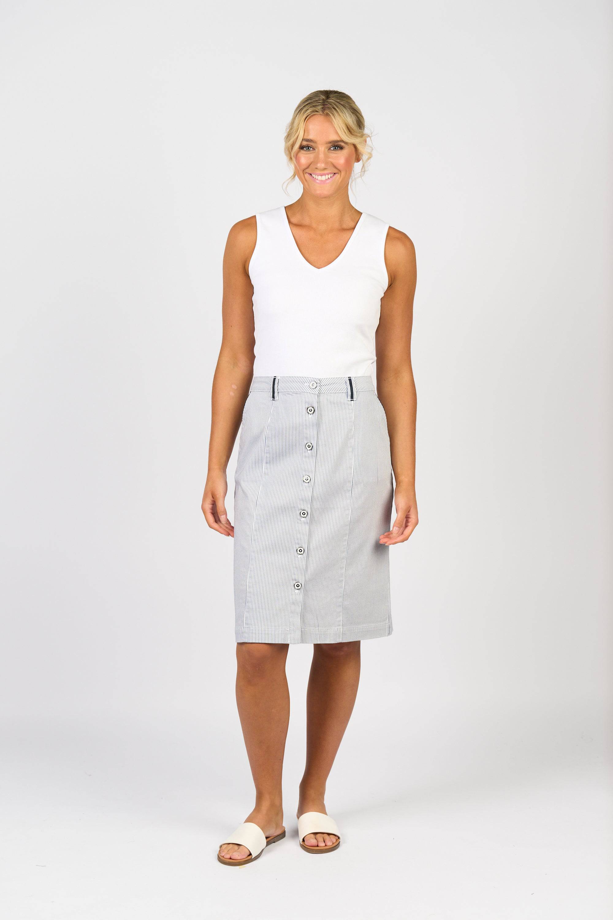 A woman with blonde hair in loose waves smiles against a white background, wearing a sleeveless white top, Vassalli's Orion Skirt - Ink/White Stripe (cotton-elastane blend, knee length with subtle stripes), and white sandals.