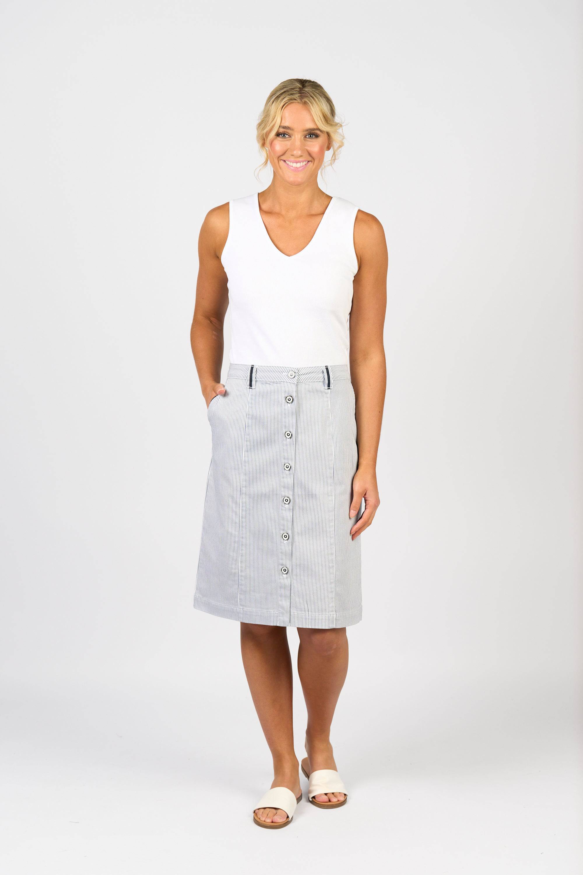A woman stands smiling in a sleeveless white top, white slide sandals, and the Vassalli Orion Skirt in Ink/White Stripe, featuring knee length and front buttons, against a plain white background.