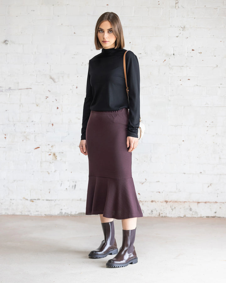 A woman in a black long-sleeve top, Vassalli’s Carrie Merino Skirt in Berry, matching boots, and a small beige shoulder bag stands against a white brick wall, looking at the camera.