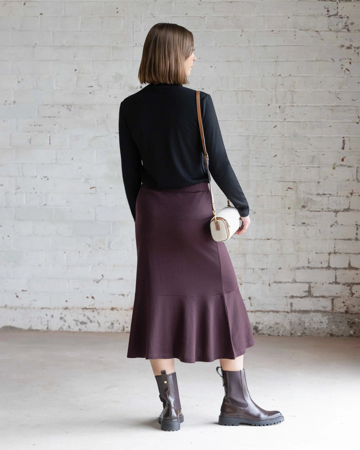 A woman with short hair stands indoors by a white brick wall, wearing a black long-sleeve top, Vassalli’s Carrie Merino Skirt in Berry with a peplum hem, brown boots, and carrying a small crossbody bag.